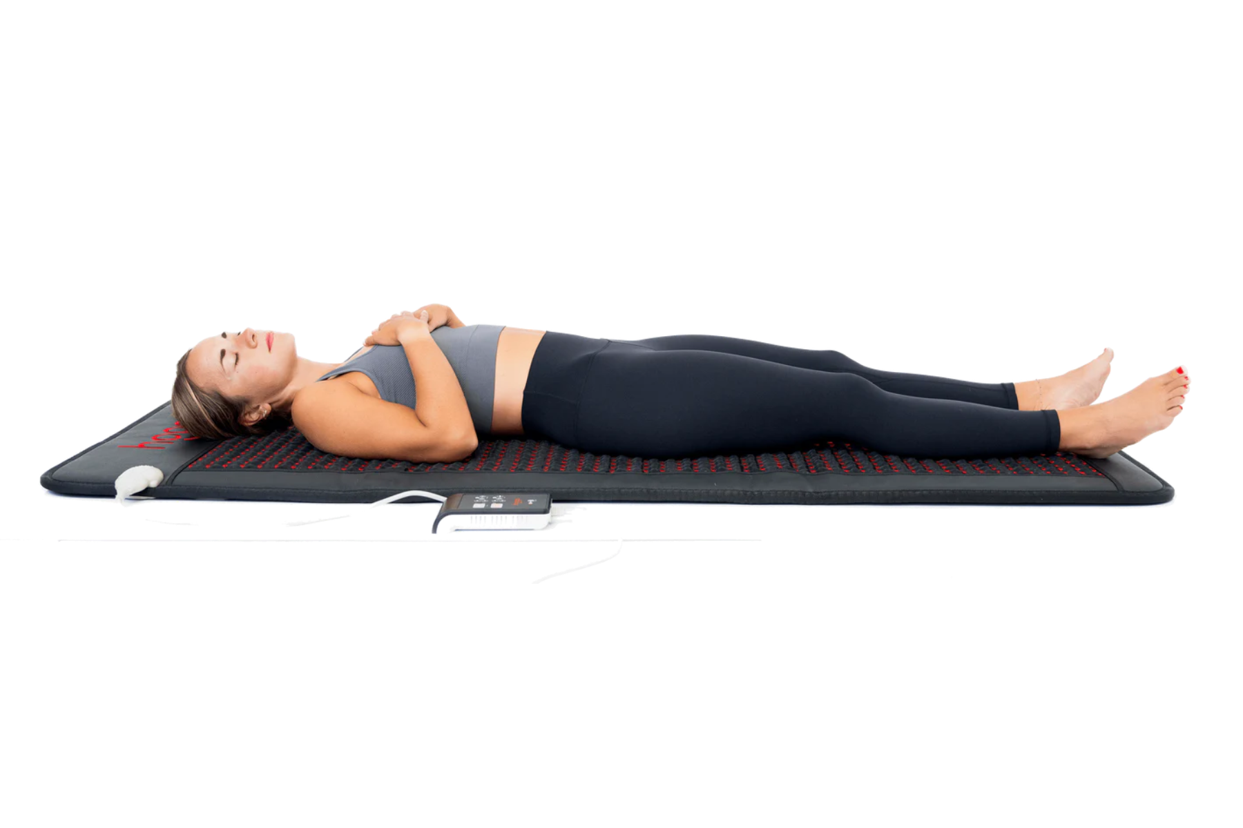 Woman relaxing on a dark gray heating mat, eyes closed, wearing gray top and black leggings.