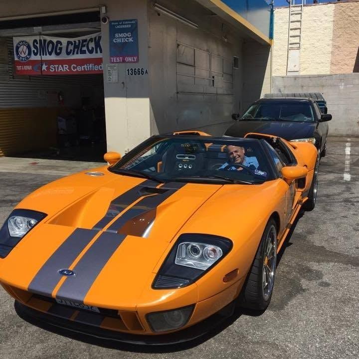 Orange Ford GT with black racing stripes parked outside a smog check shop, driver visible in the open-top car