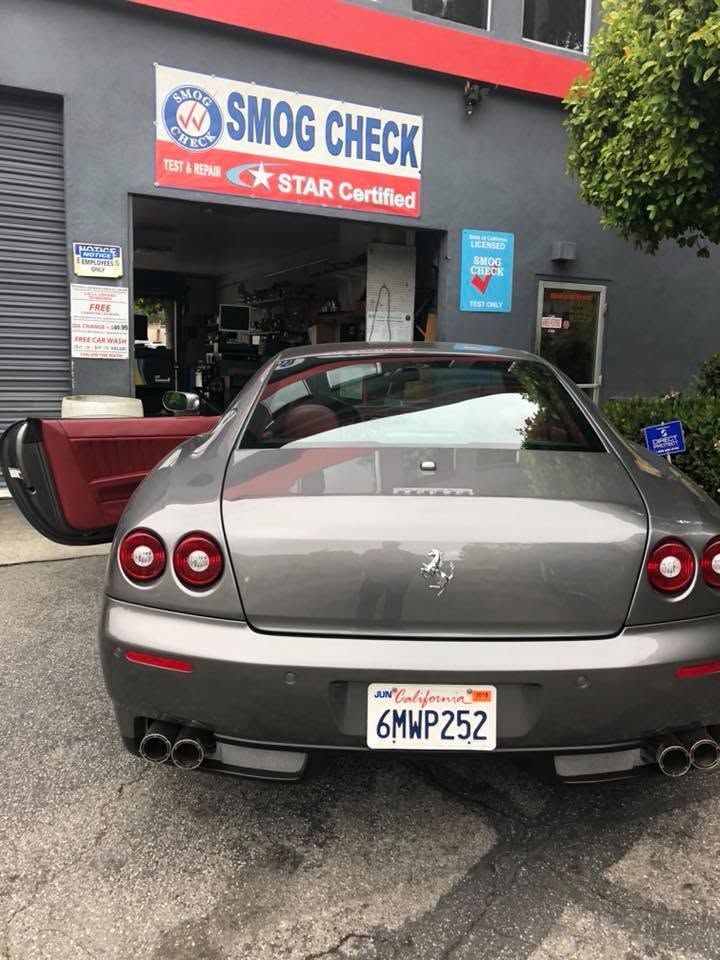 Rear view of a silver Ferrari parked at a smog check station with the driver's door open.