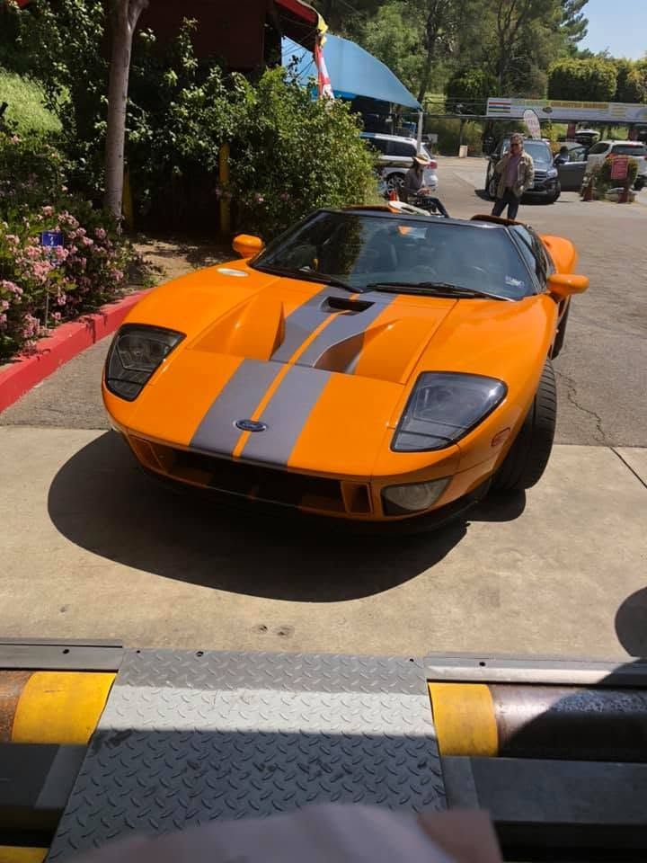 Orange sports car with gray racing stripes parked outdoors at a car show, with people and tents in the background