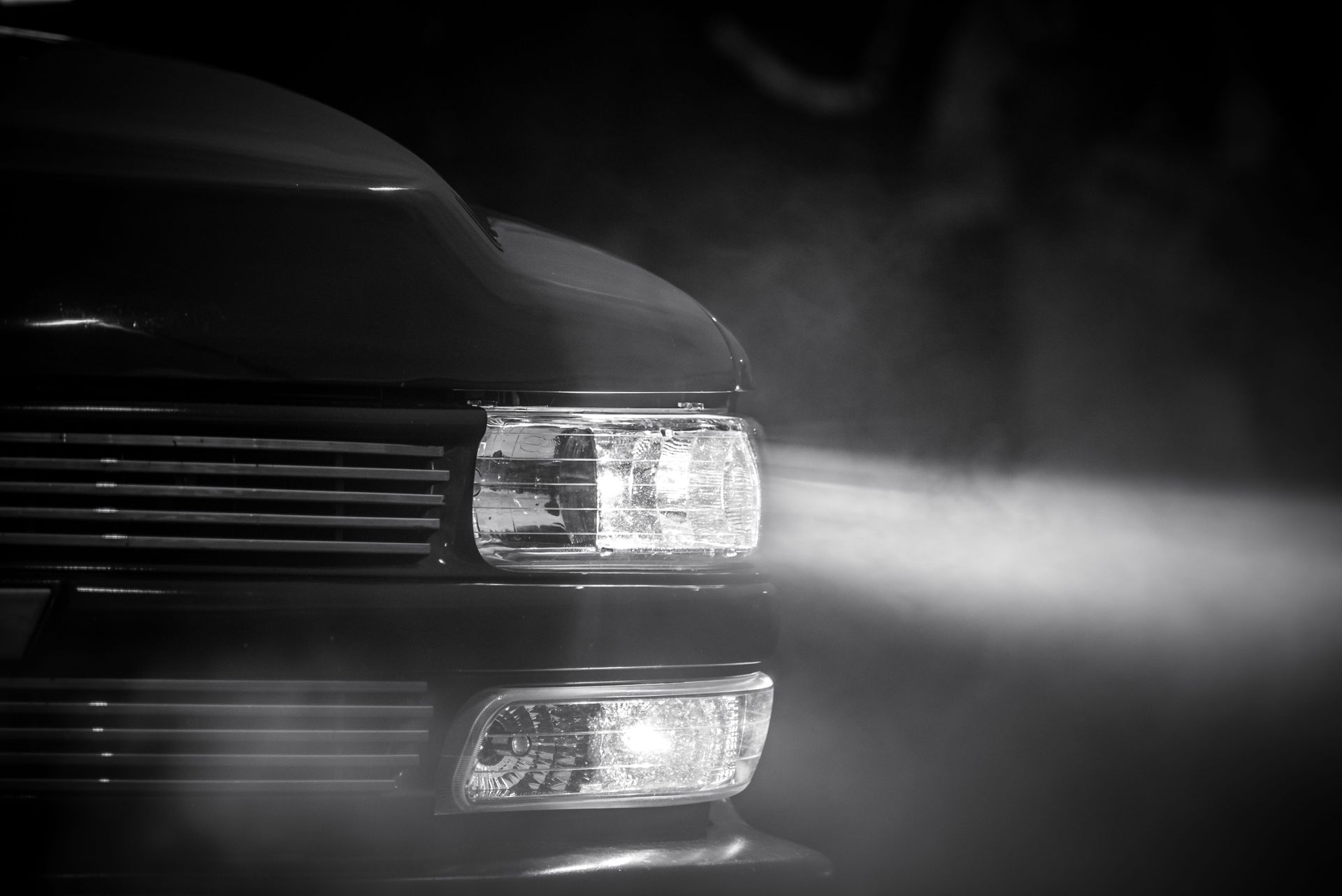 Close-up of a black car front with headlights on, shining through fog in grayscale.