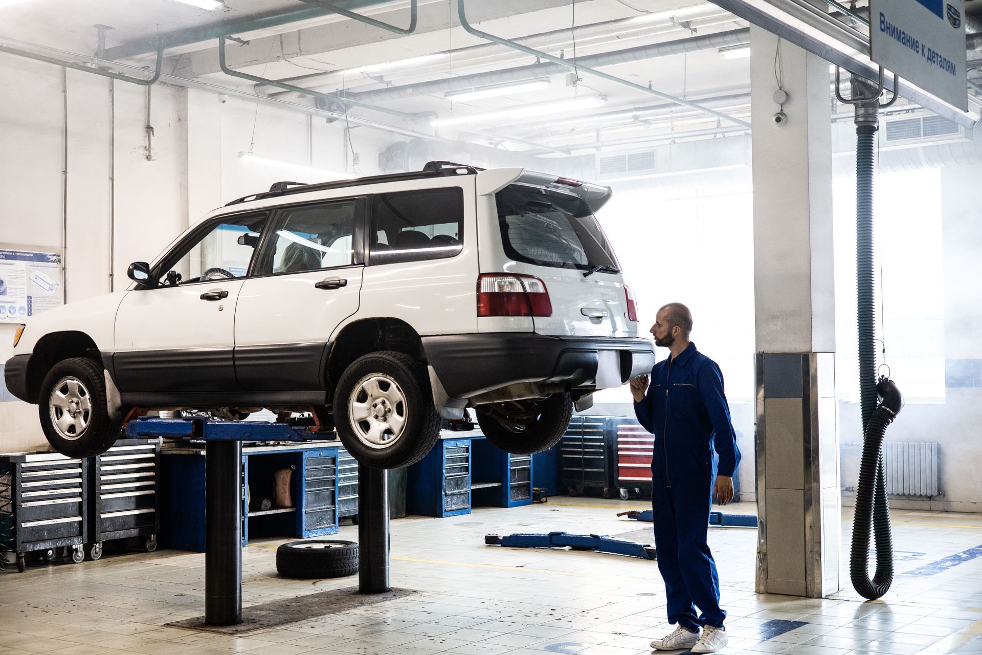 White SUV lifted on a hydraulic hoist in an auto repair shop, with a mechanic nearby