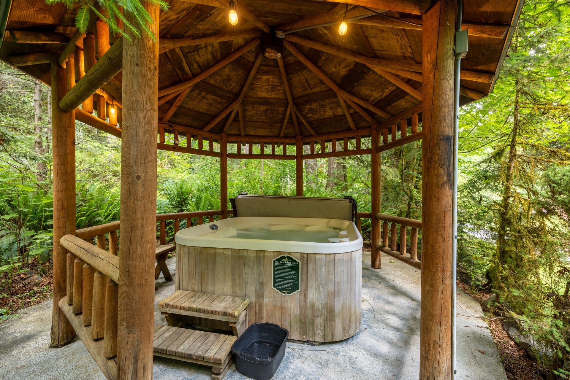 A hot tub sits inside a wooden gazebo in a forest setting. Warm lighting hangs from the ceiling.
