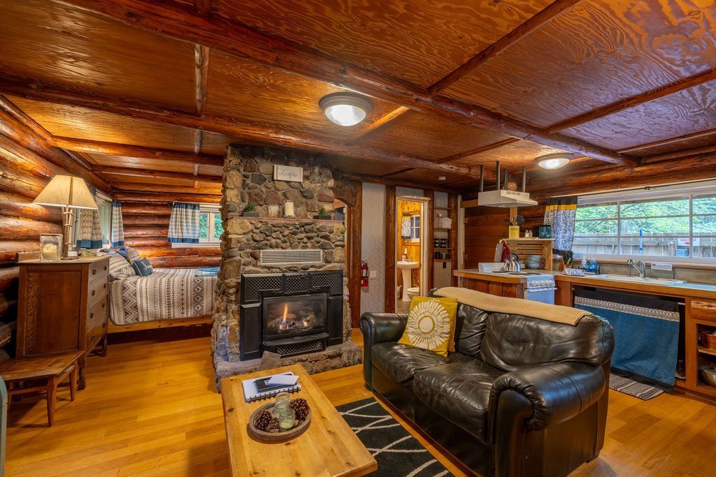 Cozy cabin living room with a stone fireplace, wooden floors, log walls, a leather sofa, and a rustic kitchen.
