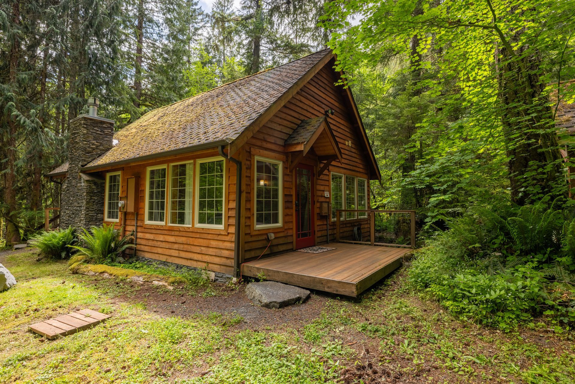 Wooden cabin in a forest setting, with a stone chimney and a small wooden deck. Surrounded by green foliage and trees.