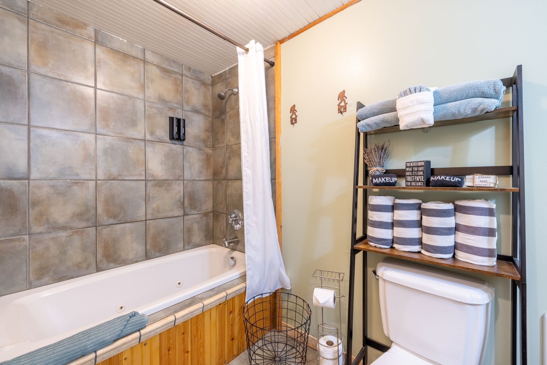 Bathroom with a bathtub, toilet, and shelving unit stocked with towels and toiletries. Light green walls, stone tile, and wood accents are present.