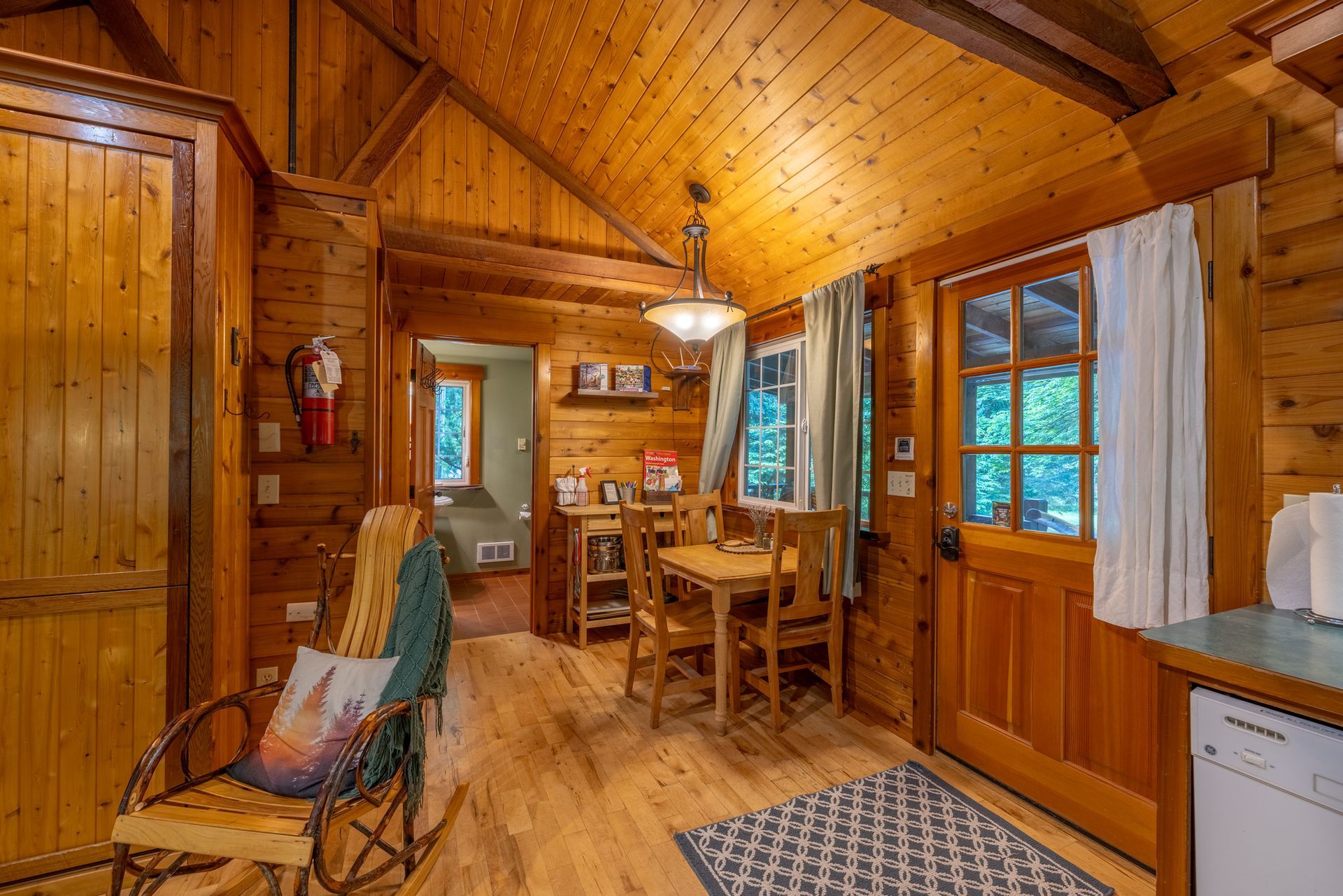 Cozy cabin interior with wooden walls and floors. A dining table, rocking chair, and doorway leading to a bedroom are visible.