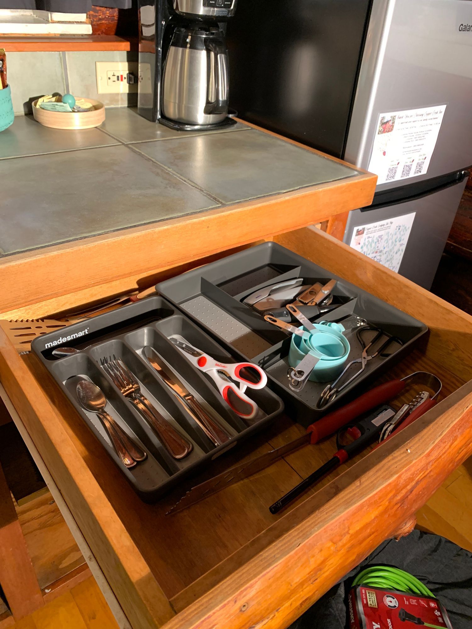 Wooden kitchen drawer with silverware and other utensils organized in gray trays. A coffee maker sits on the counter above.
