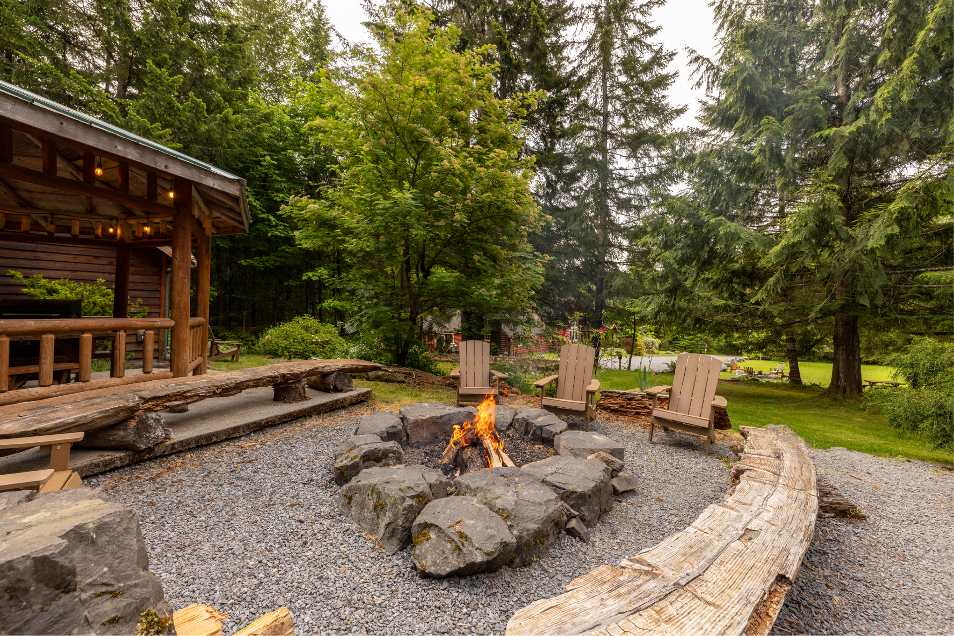 A dog sits by a campfire in a gravel-surrounded fire pit, with wooden chairs and a rustic cabin nearby in a wooded setting.