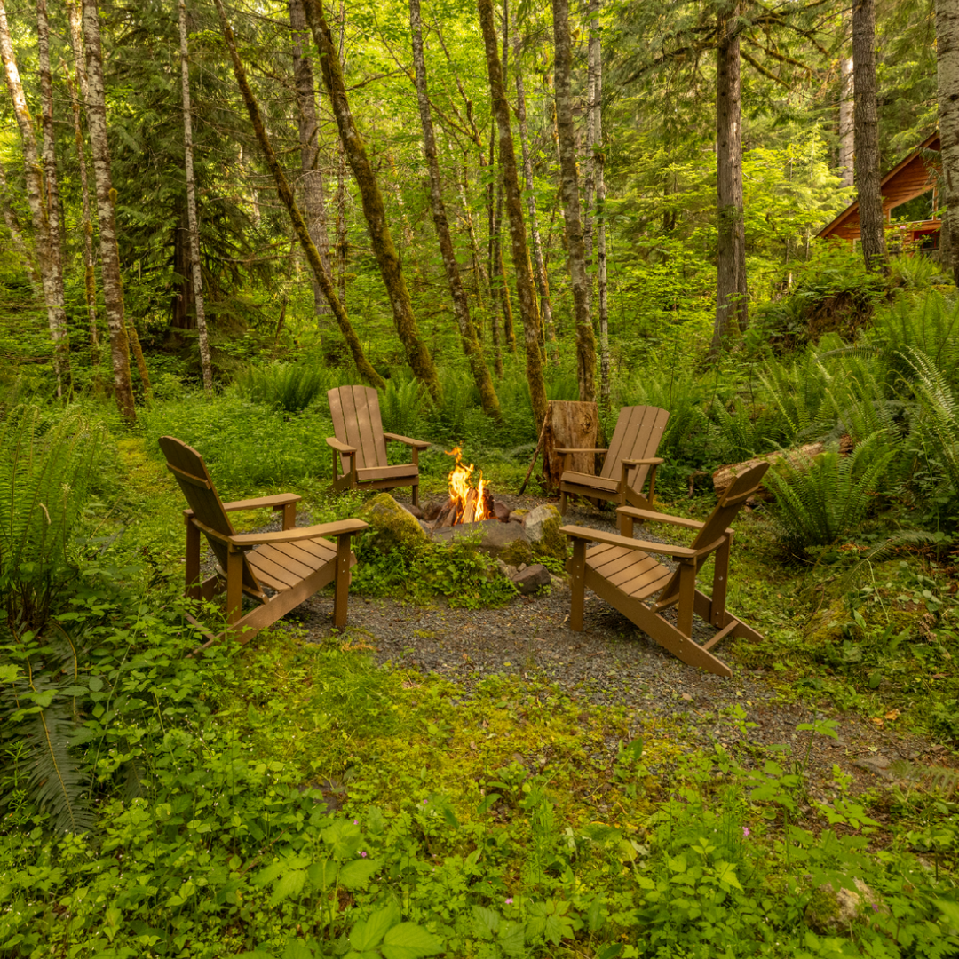 A fire pit surrounded by wooden chairs in a lush green forest setting.