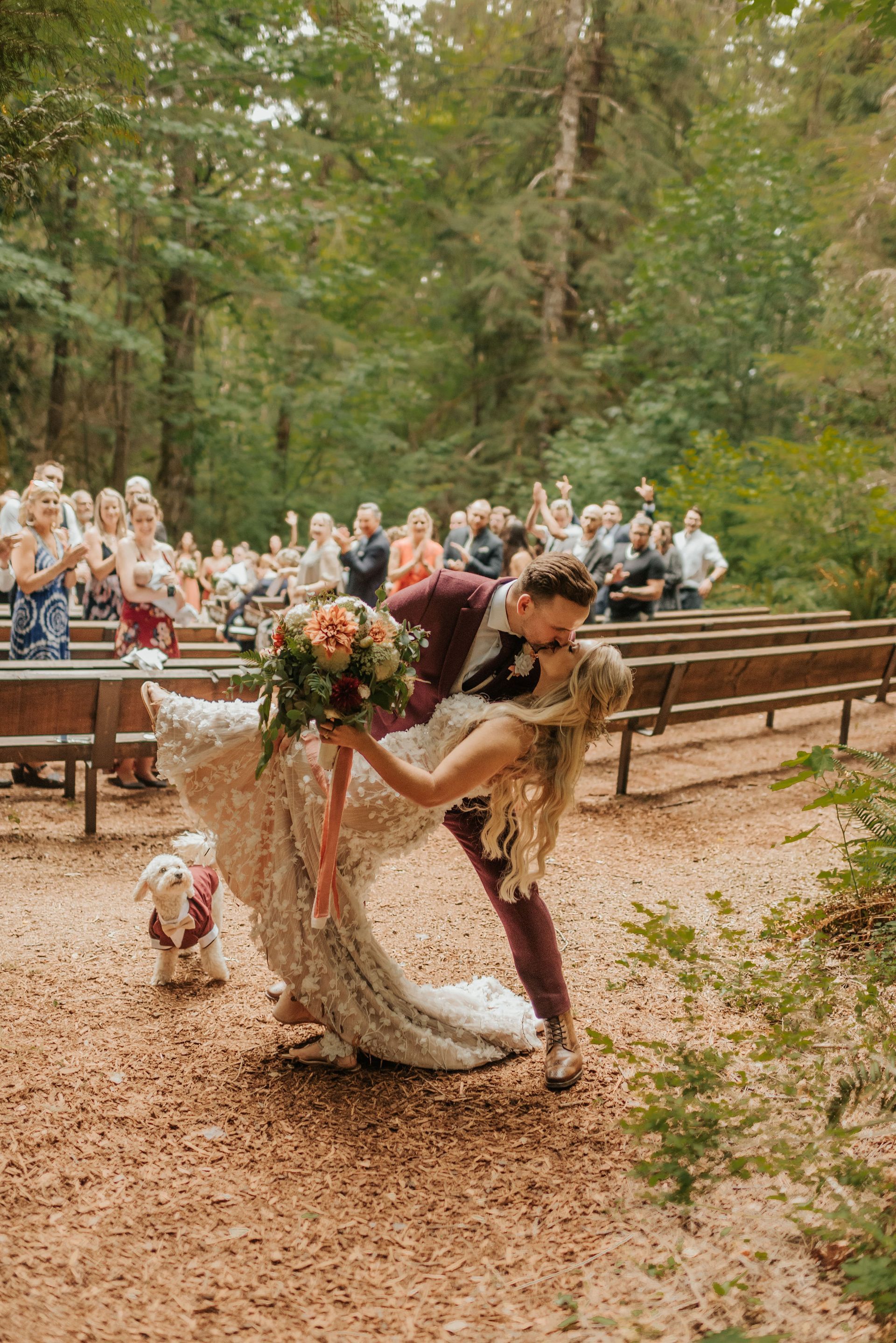 A bride and groom are kissing in front of their wedding guests while a dog watches.