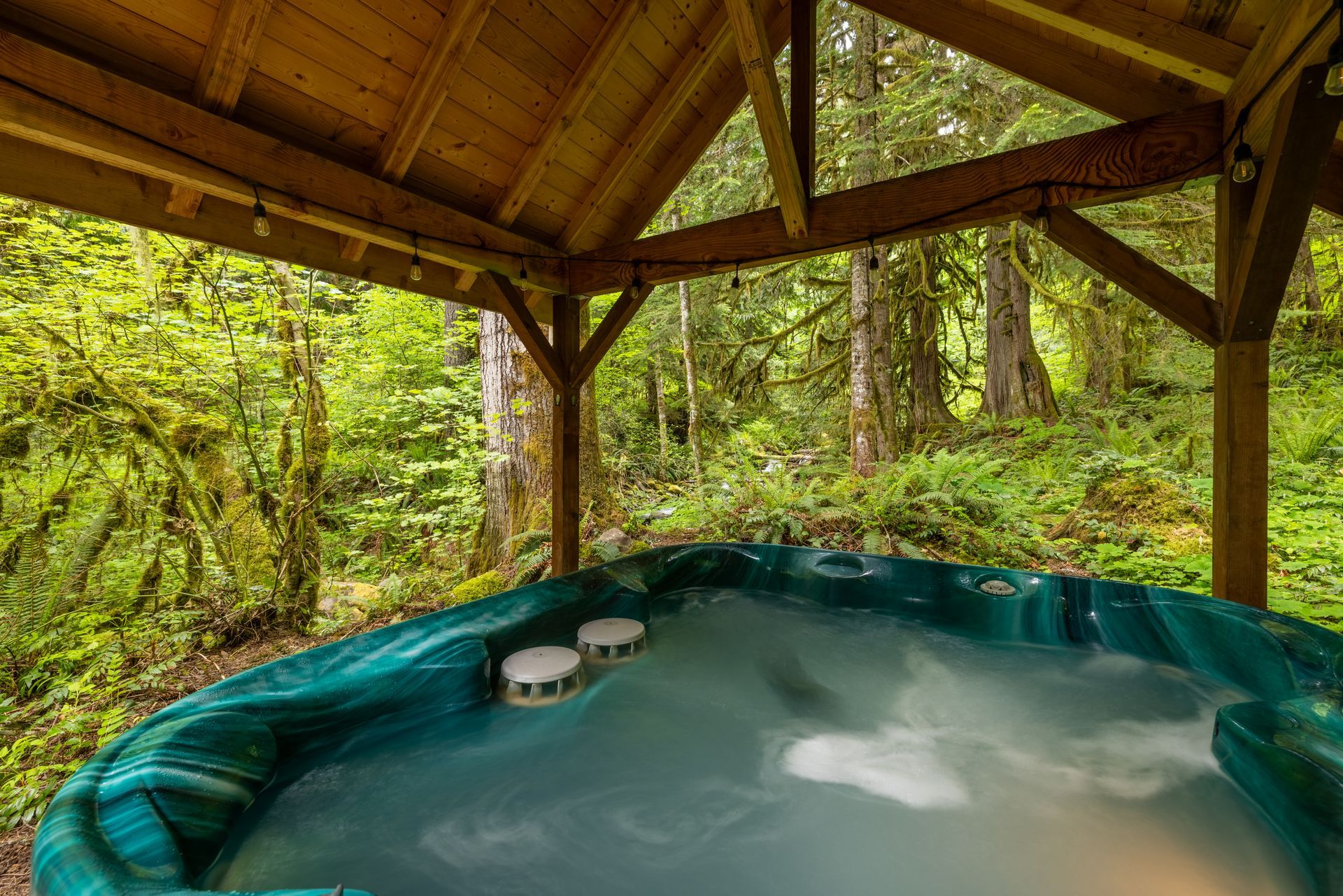 Hot tub under a wooden gazebo in a lush green forest. The water is steaming, and the setting is peaceful and secluded.