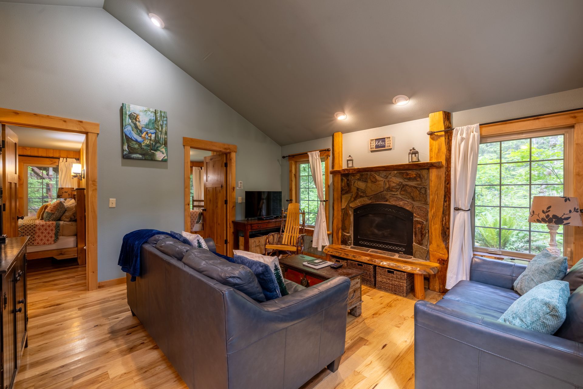 A cabin living room with a stone fireplace, wooden floors, and two blue couches. Sunlight streams in from a large window.