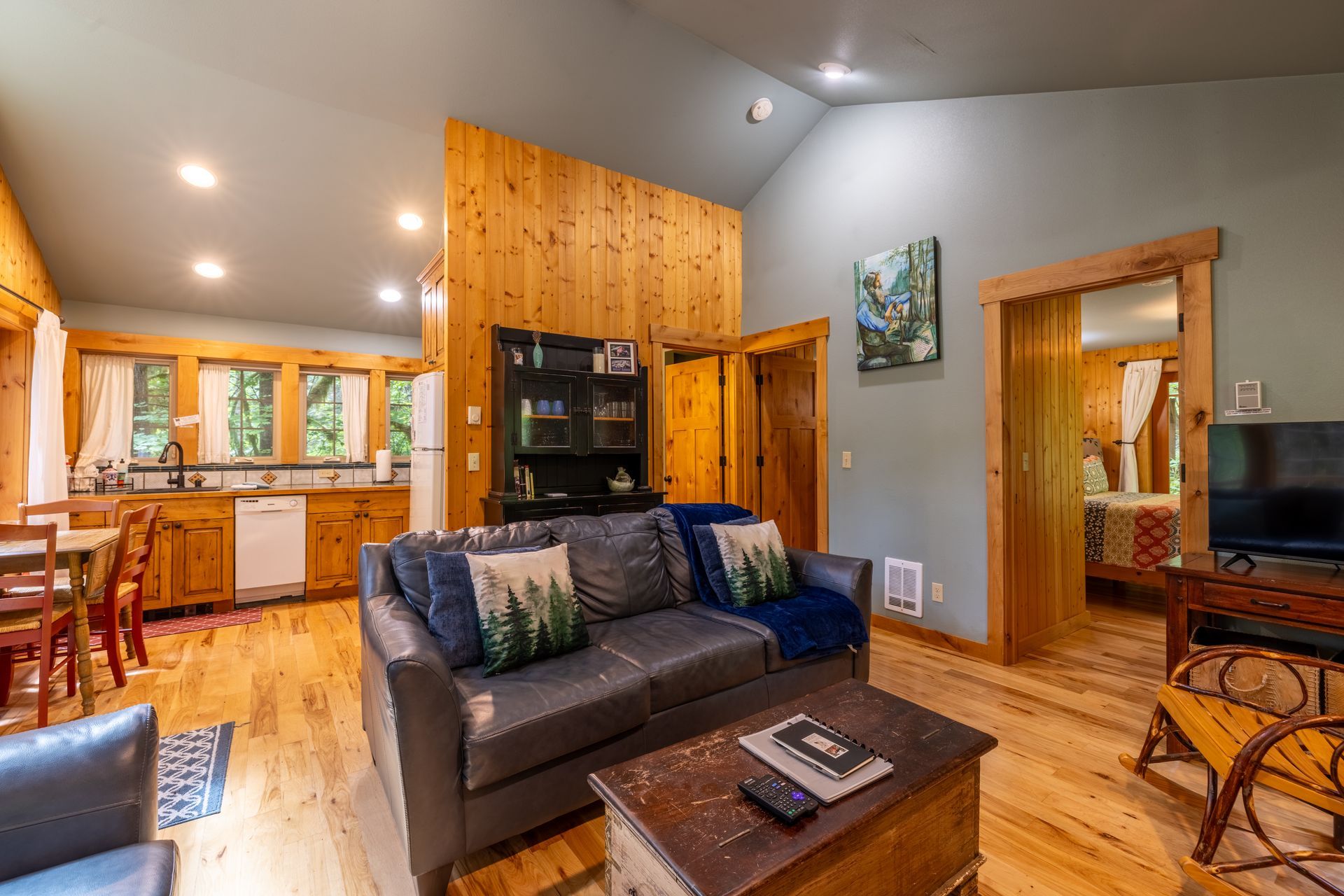 Cozy cabin interior with a living room, kitchen, and bedroom visible. Features wood floors, a gray couch, and rustic decor.