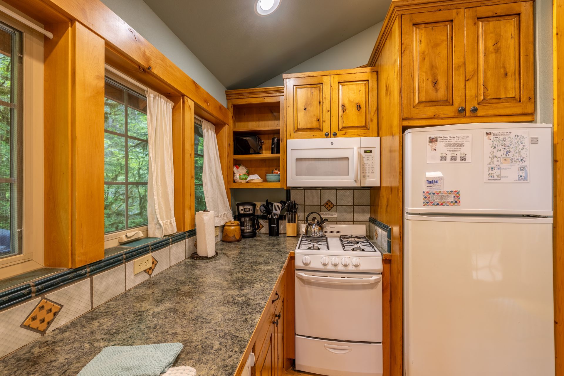 A small kitchen with wooden cabinets, a stove, and a refrigerator. A window offers a view of the outdoors.