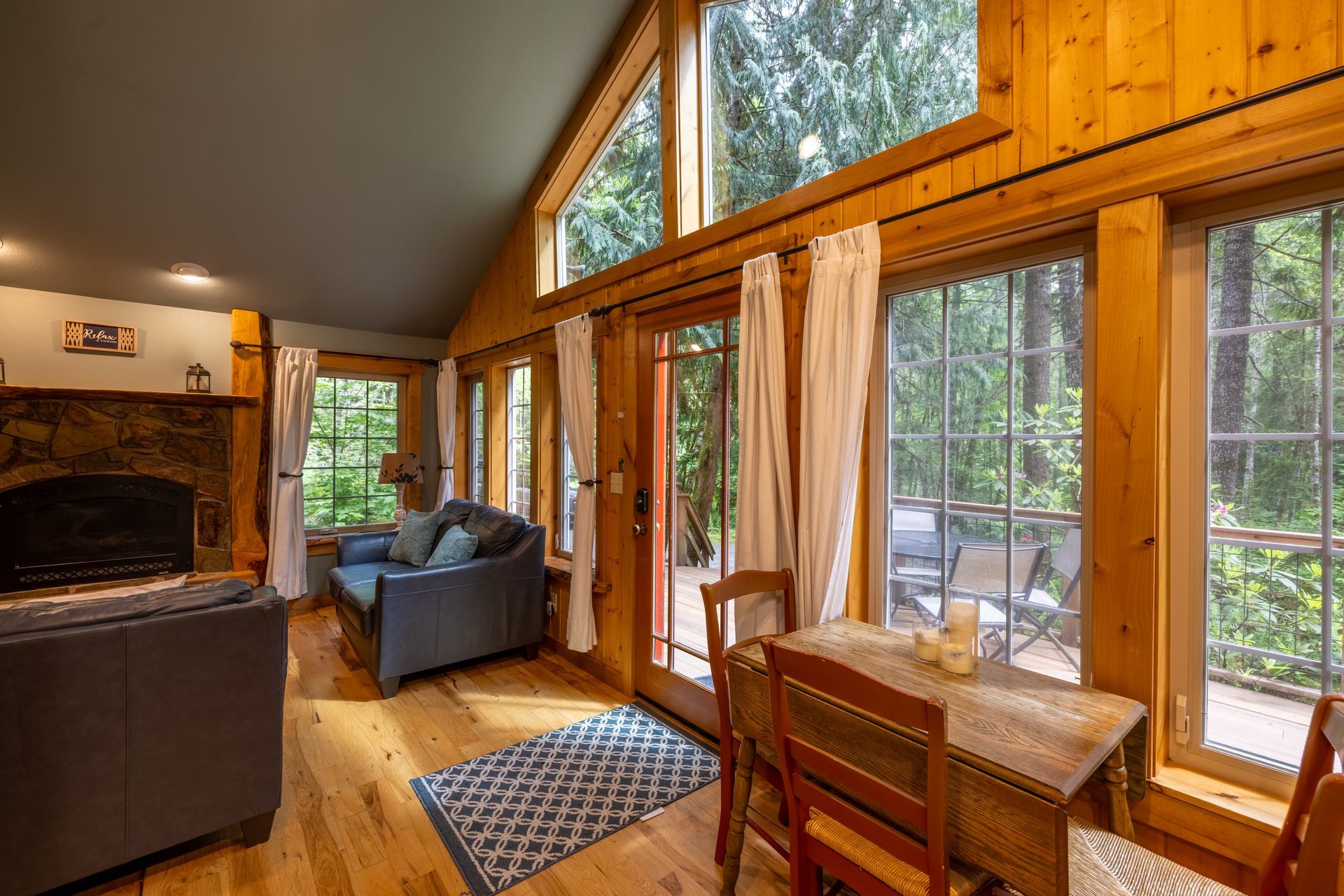 Cozy cabin living room with wooden walls, large windows overlooking trees, and a dining area. A fireplace and blue sofa are visible.