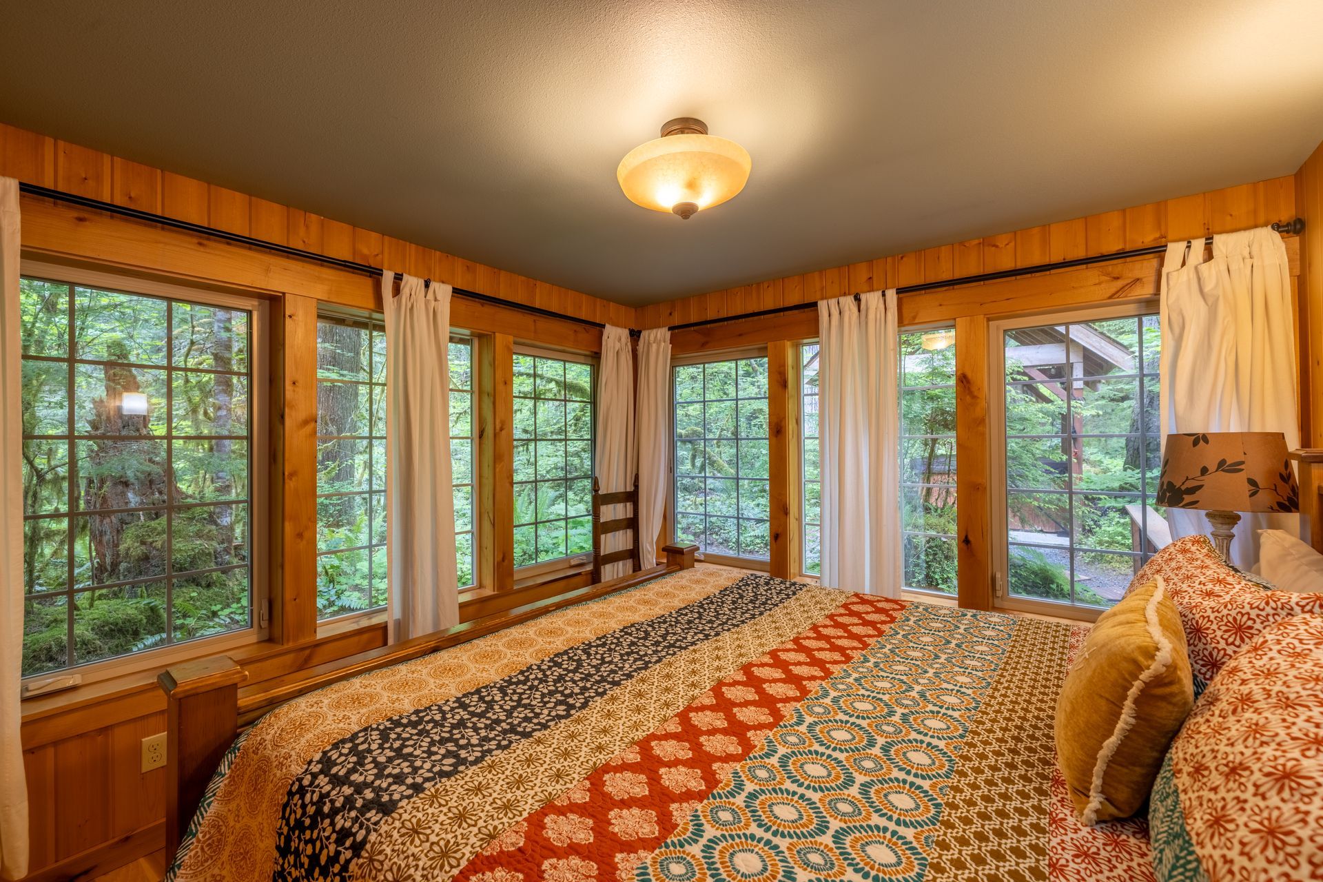 Bedroom with large windows overlooking a lush green forest. A bed with a patterned quilt and pillows is in the foreground.