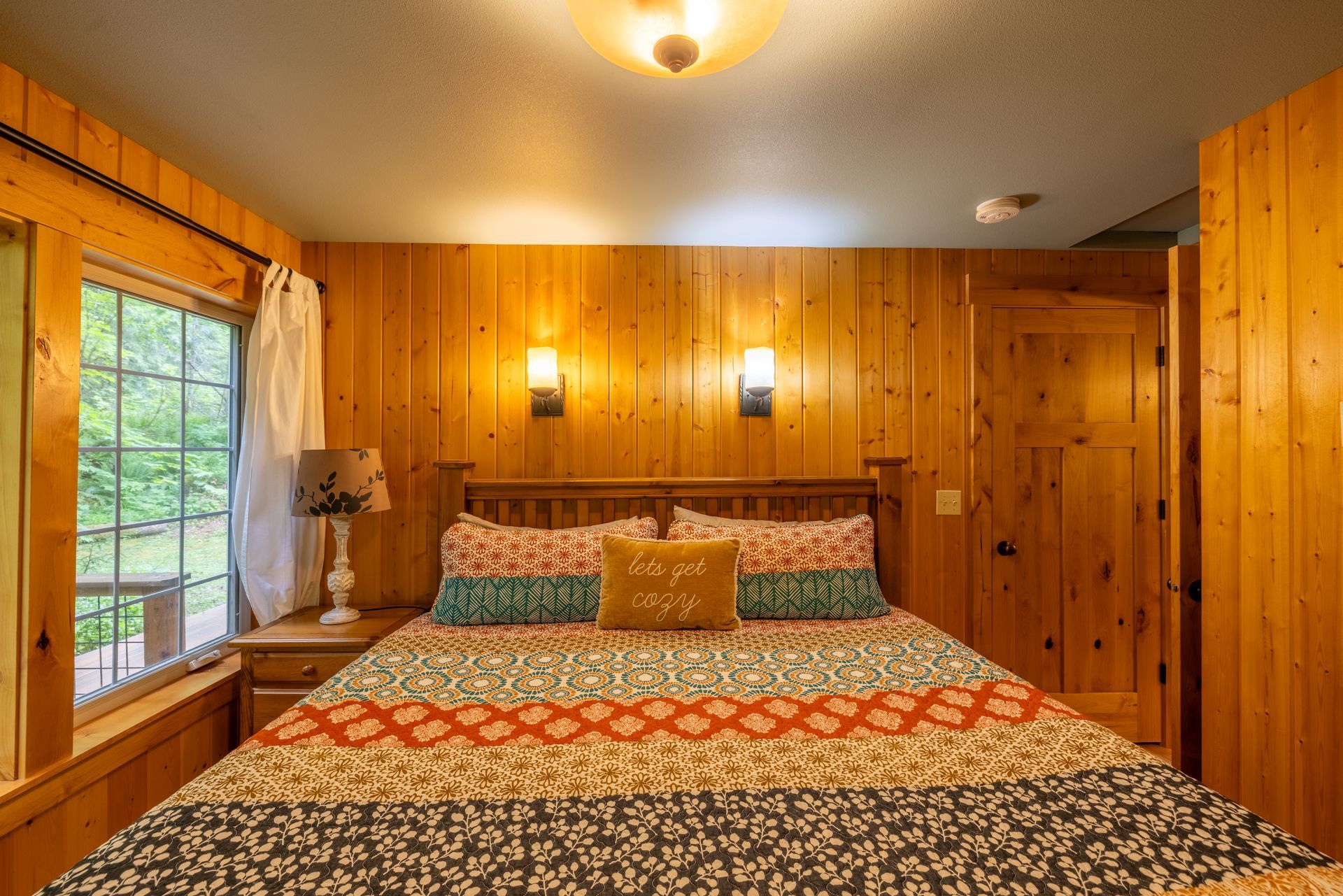 Bedroom with wooden walls and a large bed with colorful bedding. A window with a view and two sconces are visible.
