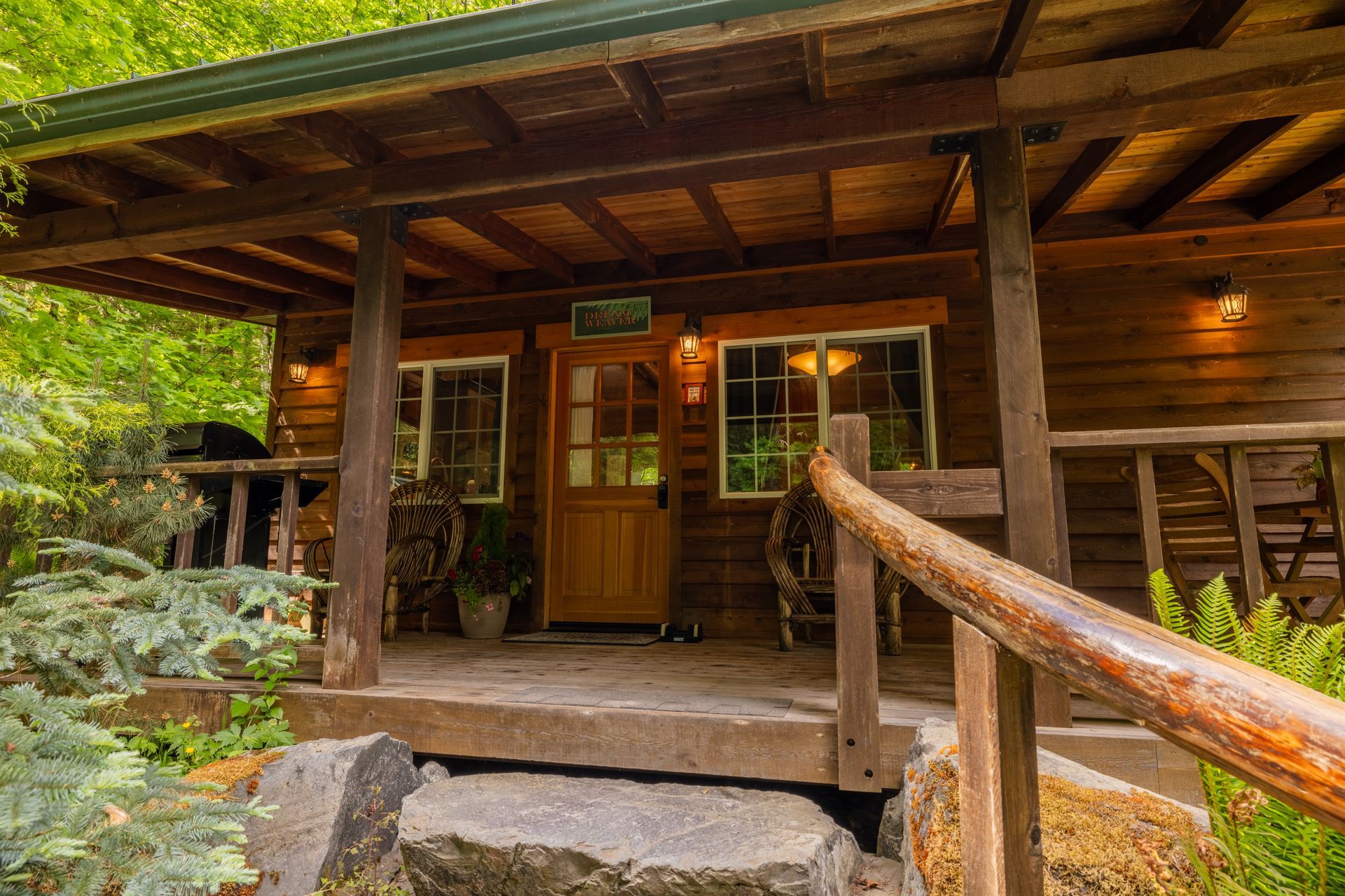 Wooden cabin with a porch and steps leading up. Green foliage surrounds the structure; warm lighting.
