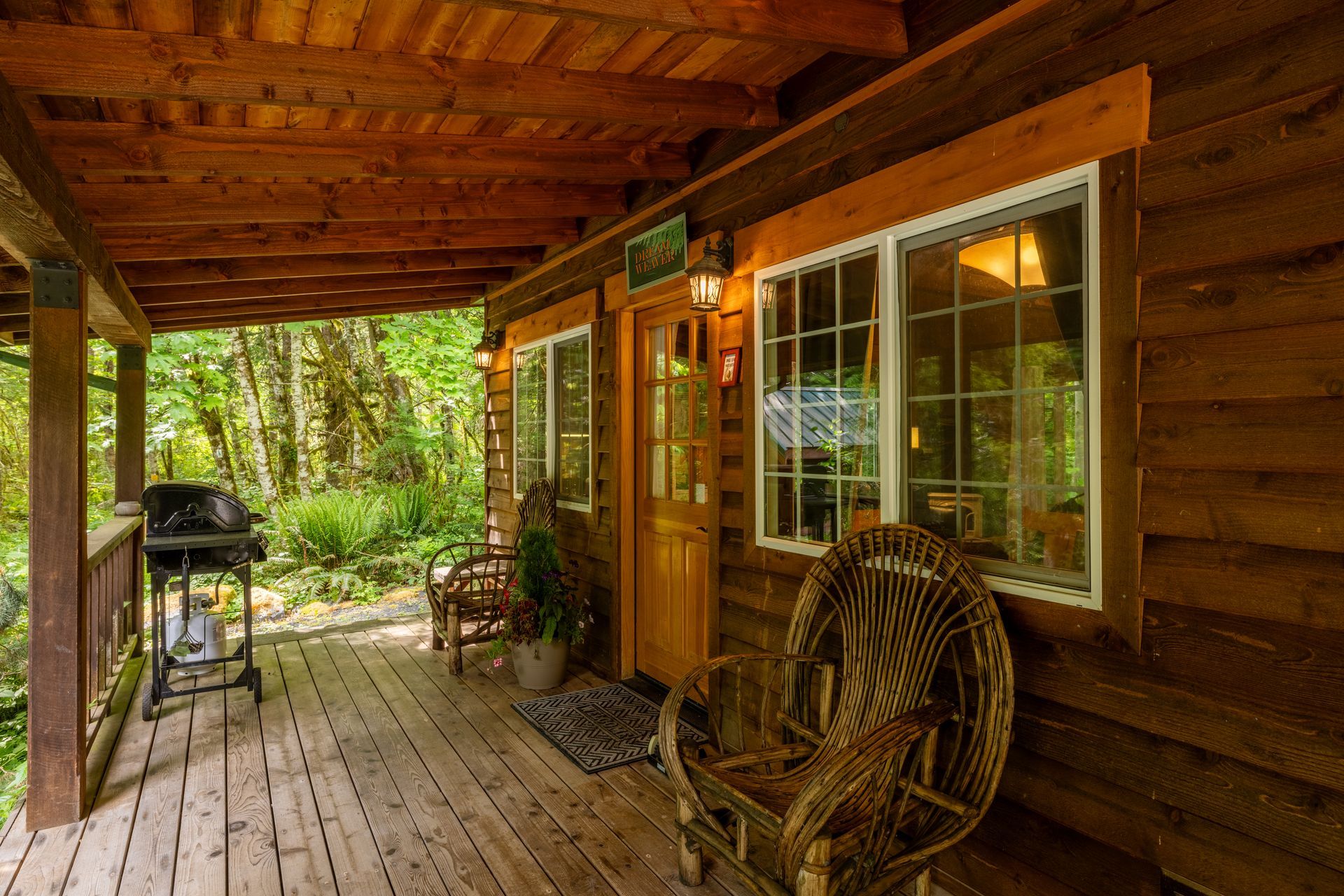 Wooden cabin porch with a grill, wicker chair, and two windows overlooking a forested area.