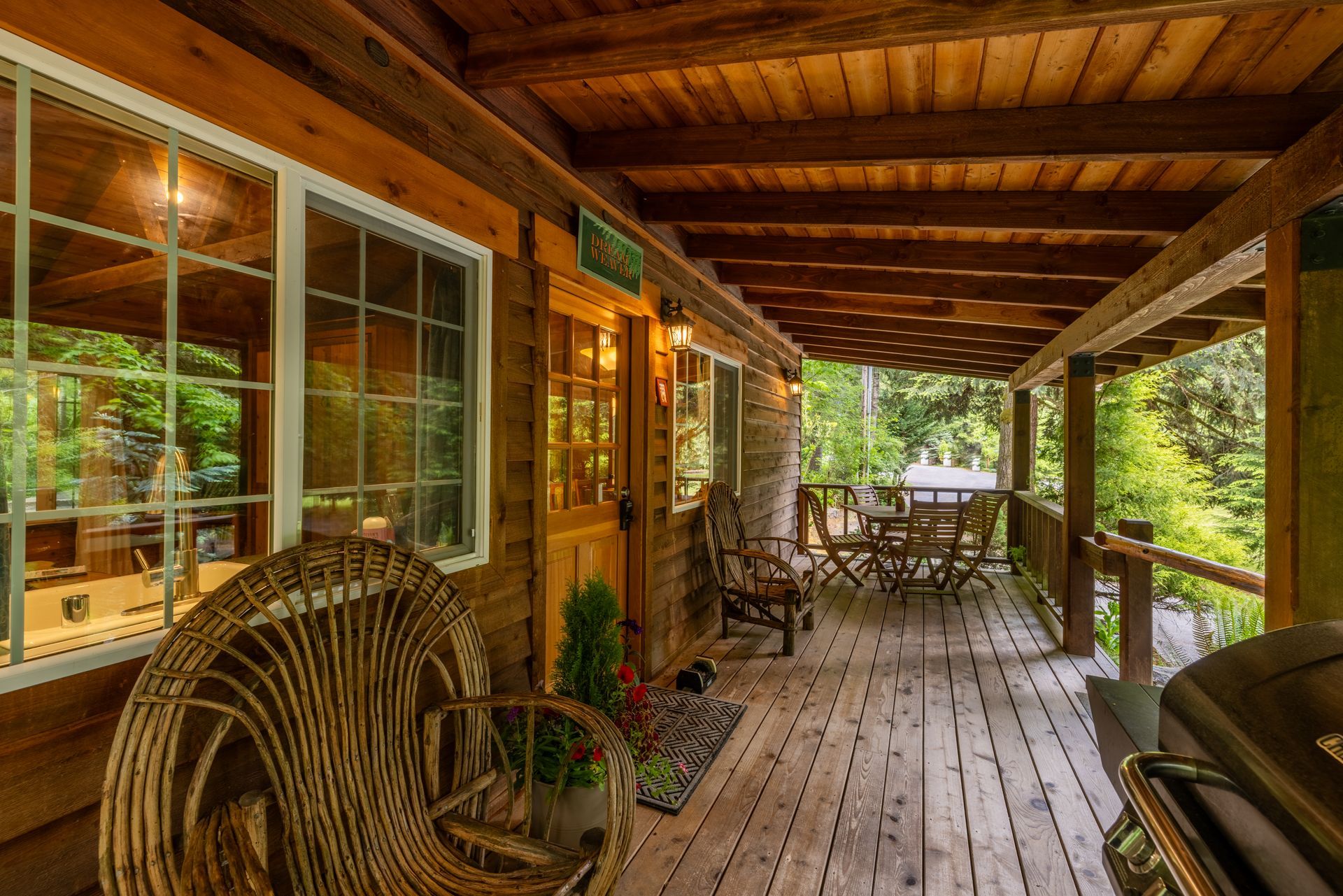 Wooden cabin porch with wicker chair, table and chairs, and lush greenery visible. Warm, inviting atmosphere.