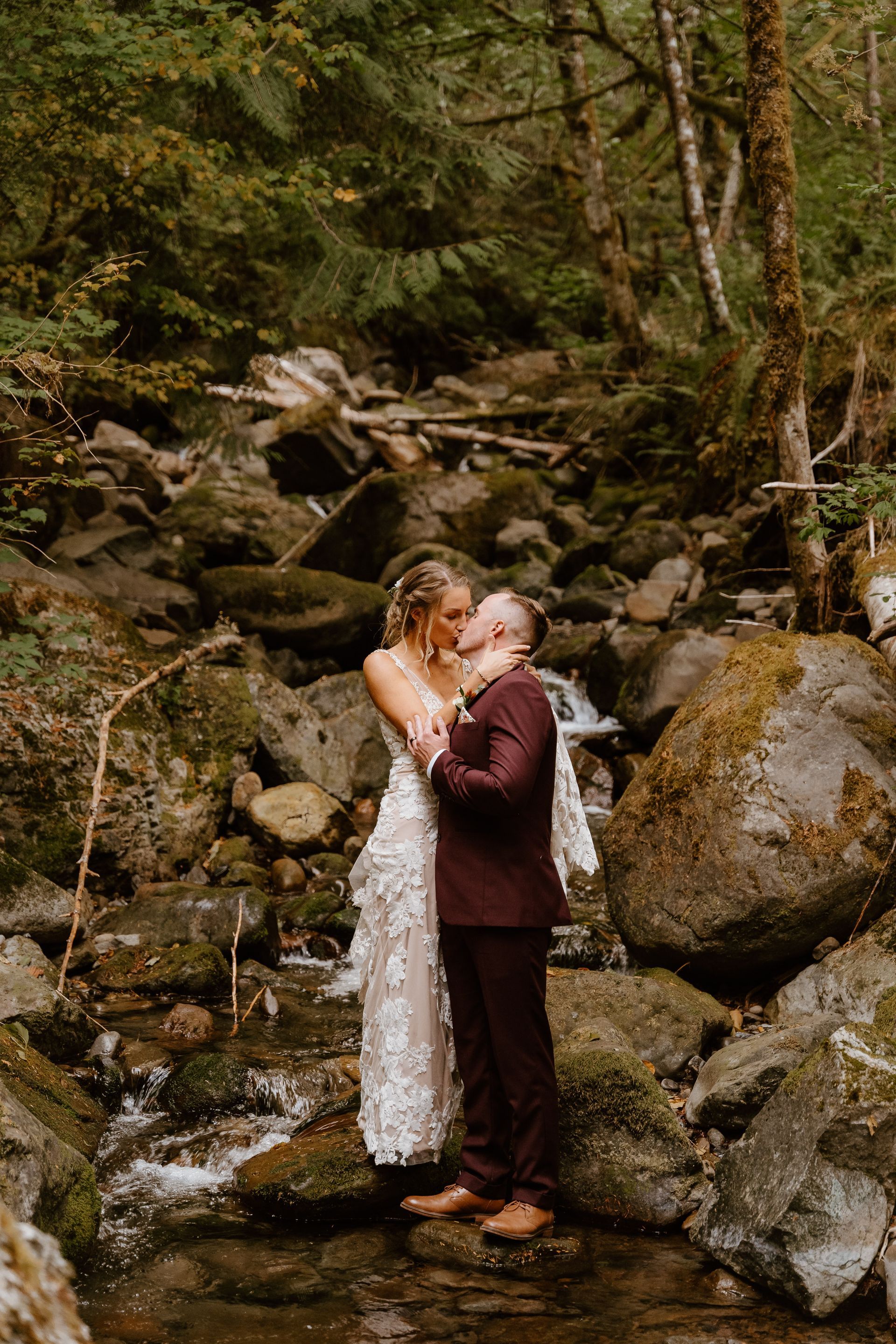 A bride and groom are kissing in a stream in the woods.
