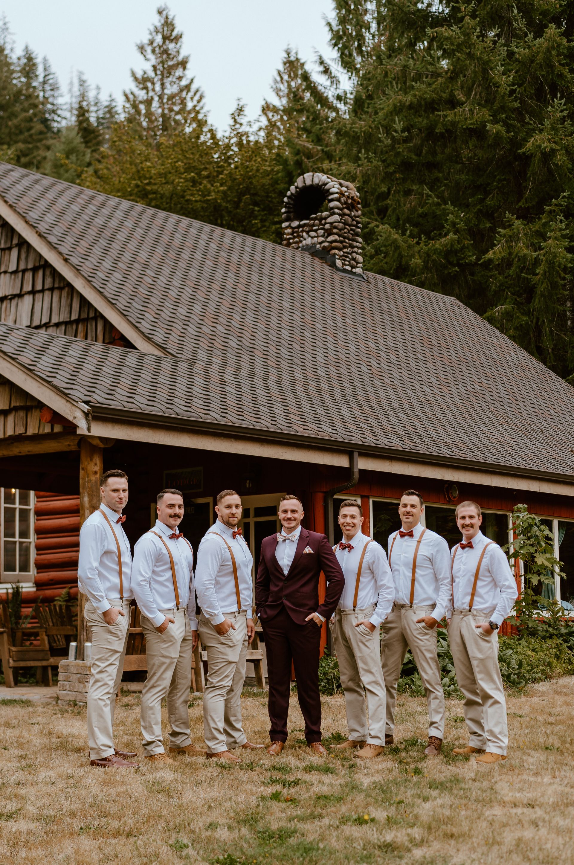 The groom and his groomsmen are posing for a picture in front of a log cabin.