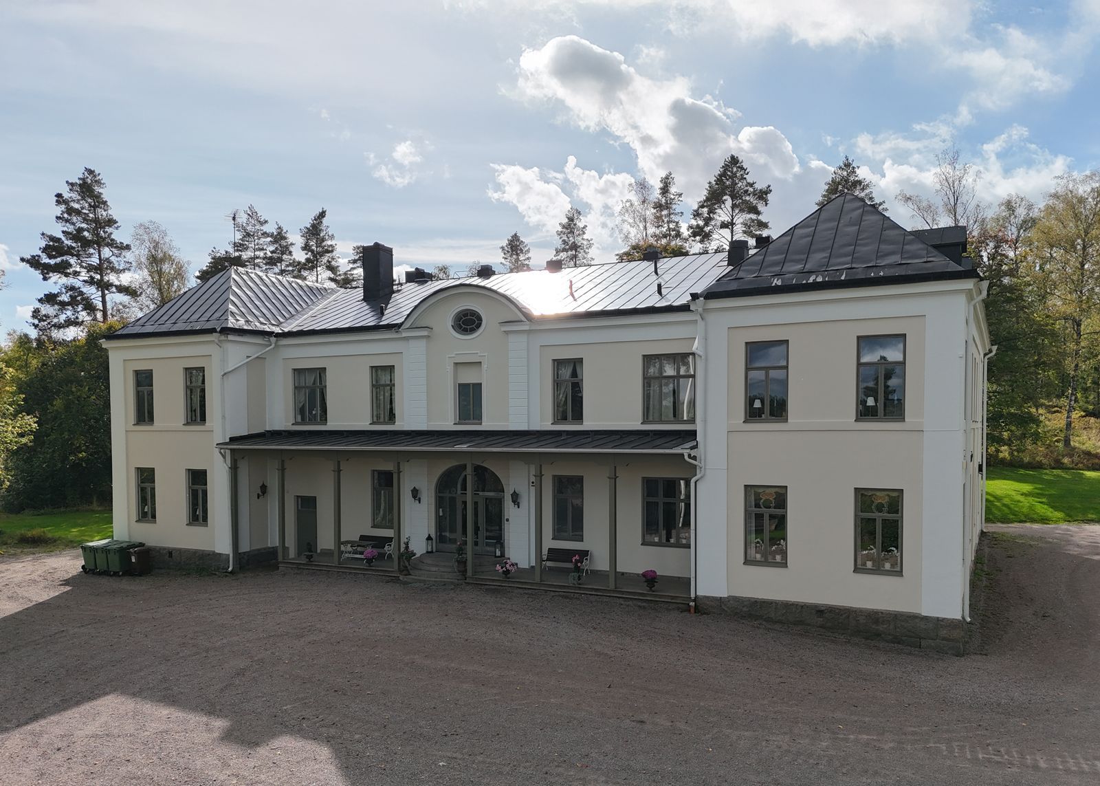 Stort vitt herrgård med pelare och mörkt tak, beläget bredvid en grusuppfart under en molnig himmel