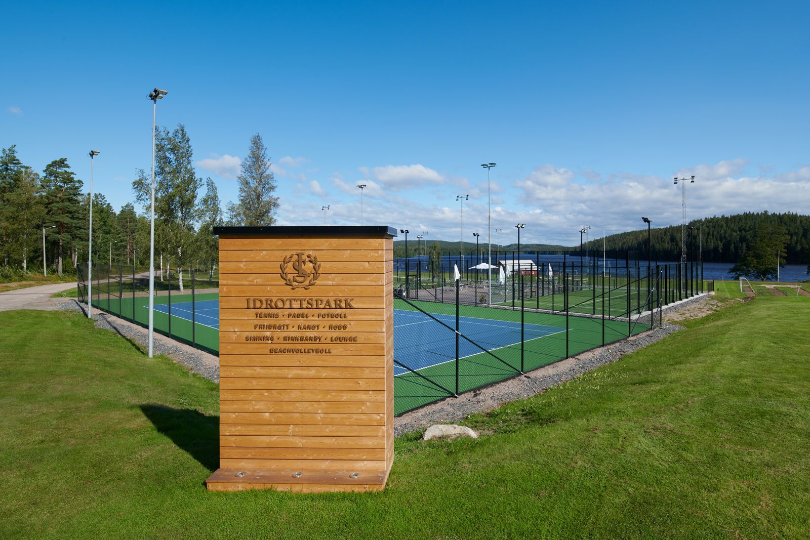 Tennisbanor bakom en träskylt i en gräsbevuxen park under en blå himmel