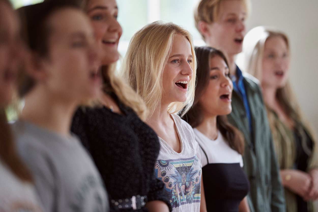 Människor som sjunger i rad inomhus, med en blond kvinna i mitten och andra suddiga i bakgrunden.