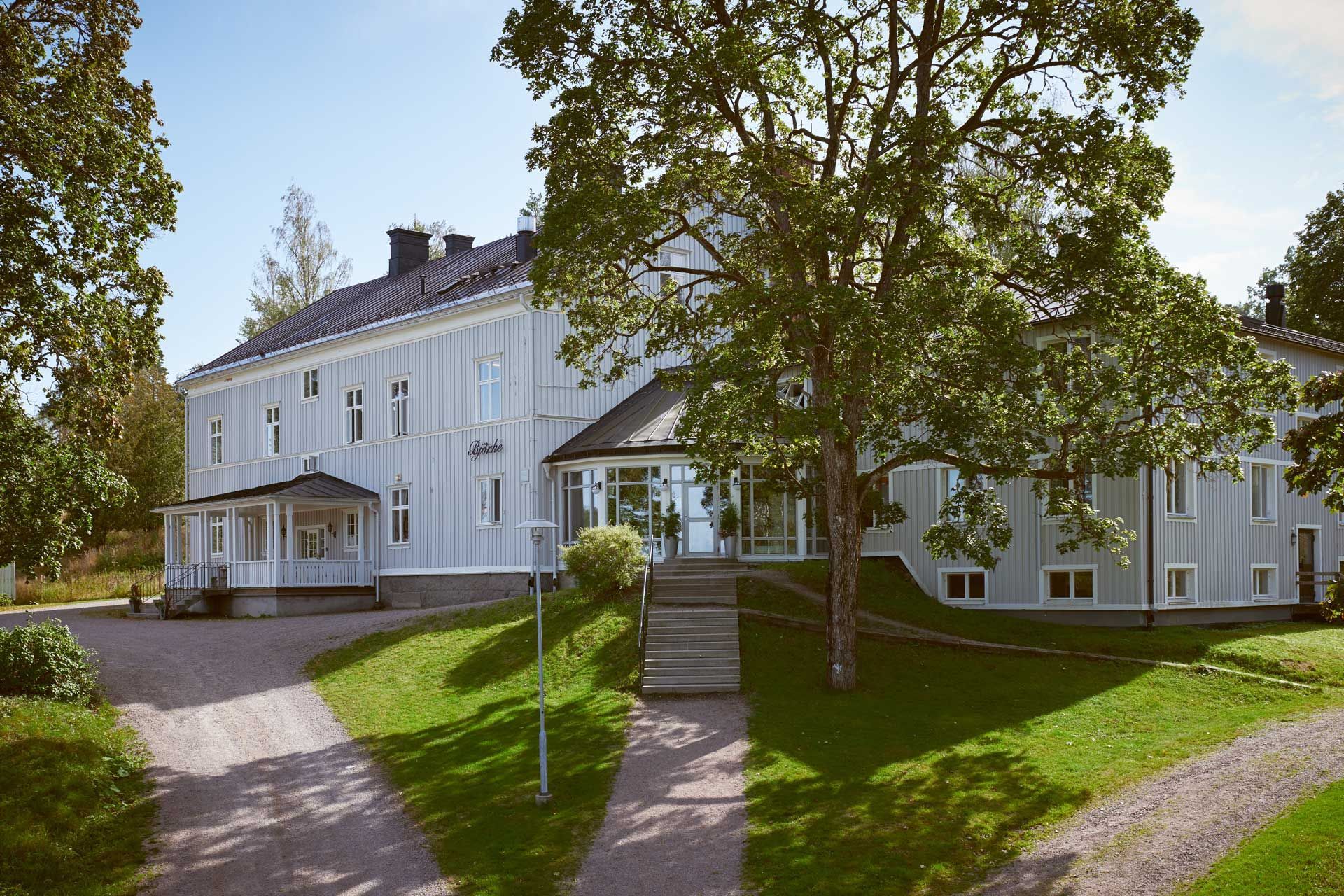 Vitt tvåvåningshus med veranda och stora träd, beläget på en gräsmatta med en stenstig och trappor