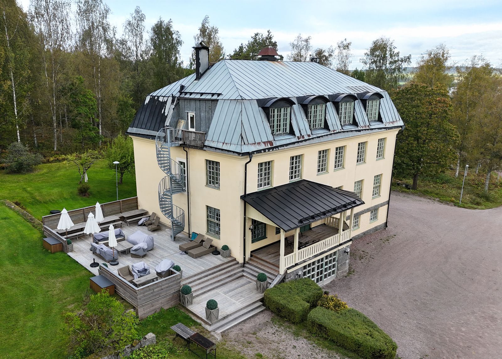 Flygfoto över ett stort beige hus med grått tak, uteplats och omgivande träd.