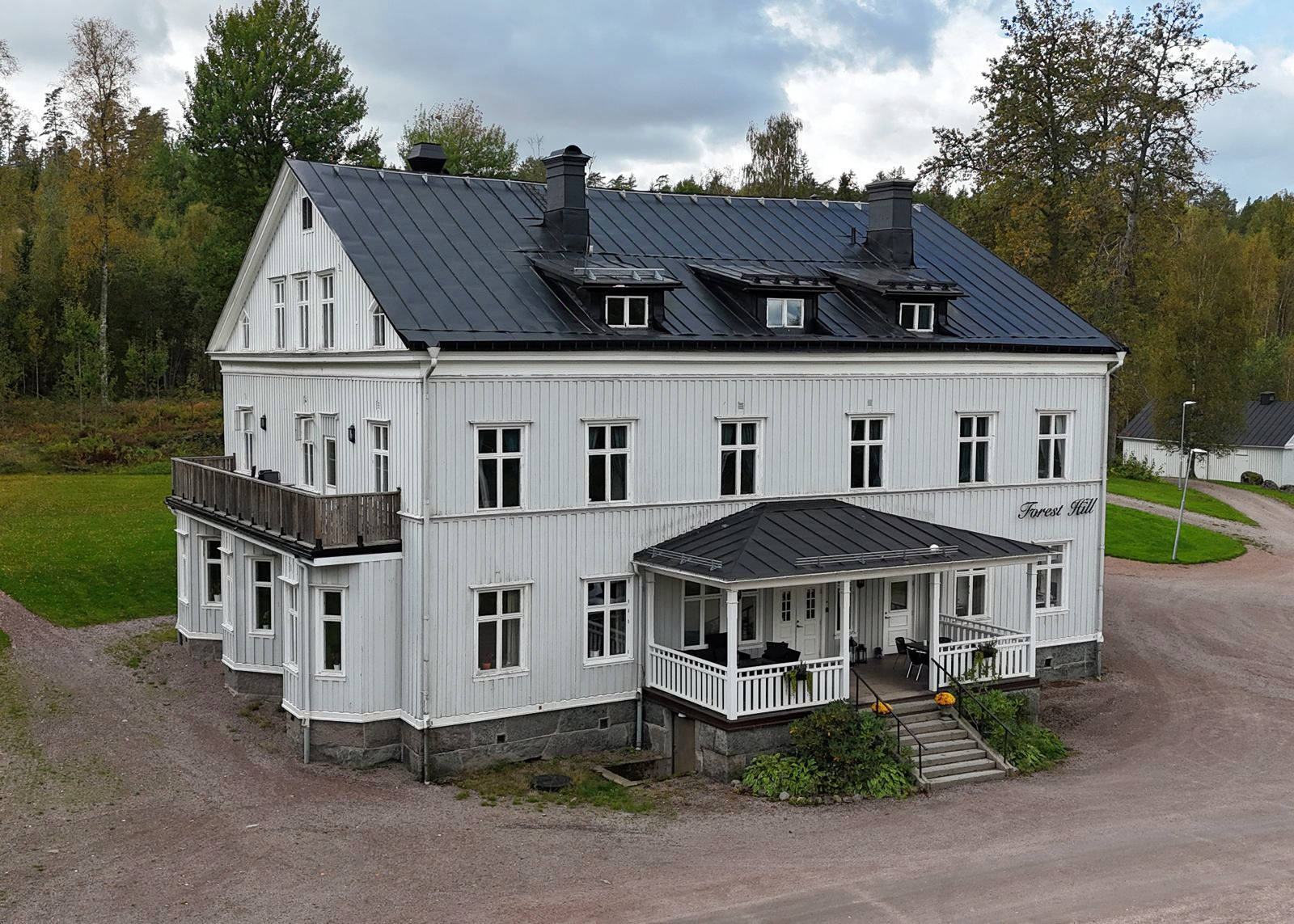 Stort vitt tvåvåningshus med mörkt tak och balkonger, omgivet av träd och grusuppfart