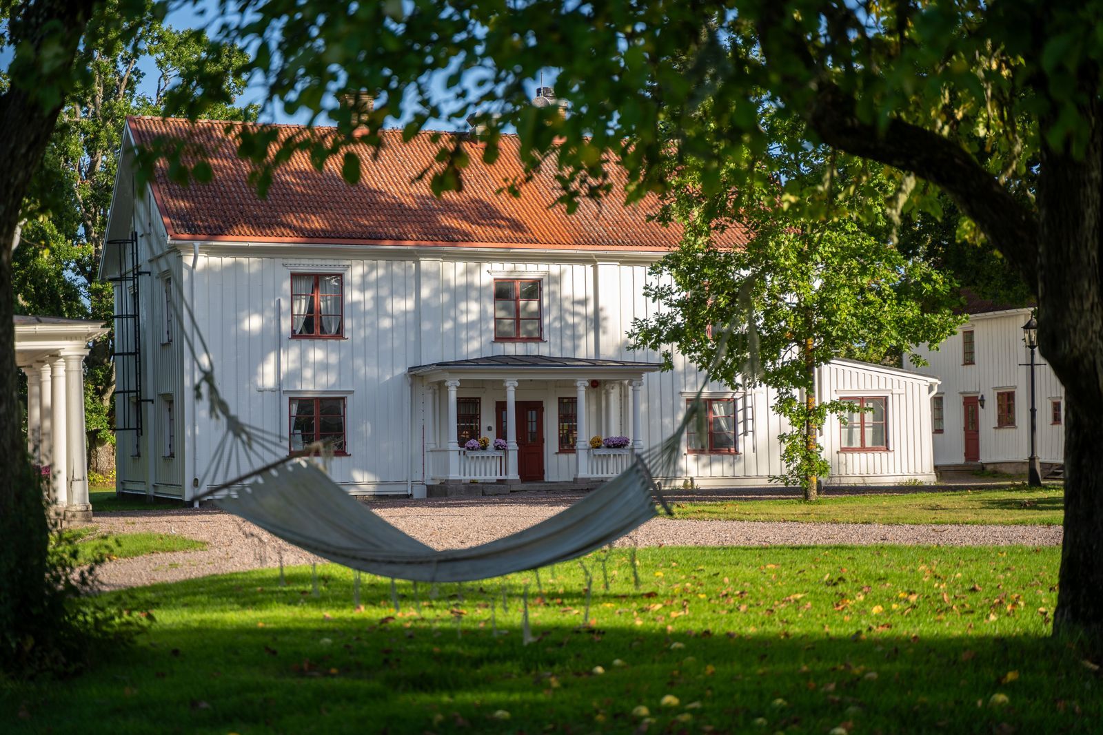 Vitt hus med rött tak och hängmatta i en skuggig grön gård