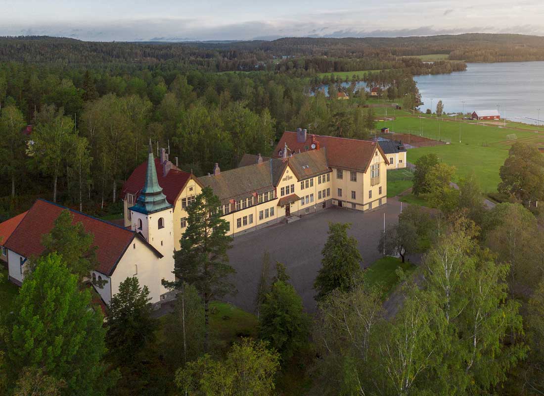 Flygfoto över en historisk slottsliknande byggnad med röda tak bredvid en sjö och skogsklädda marker.