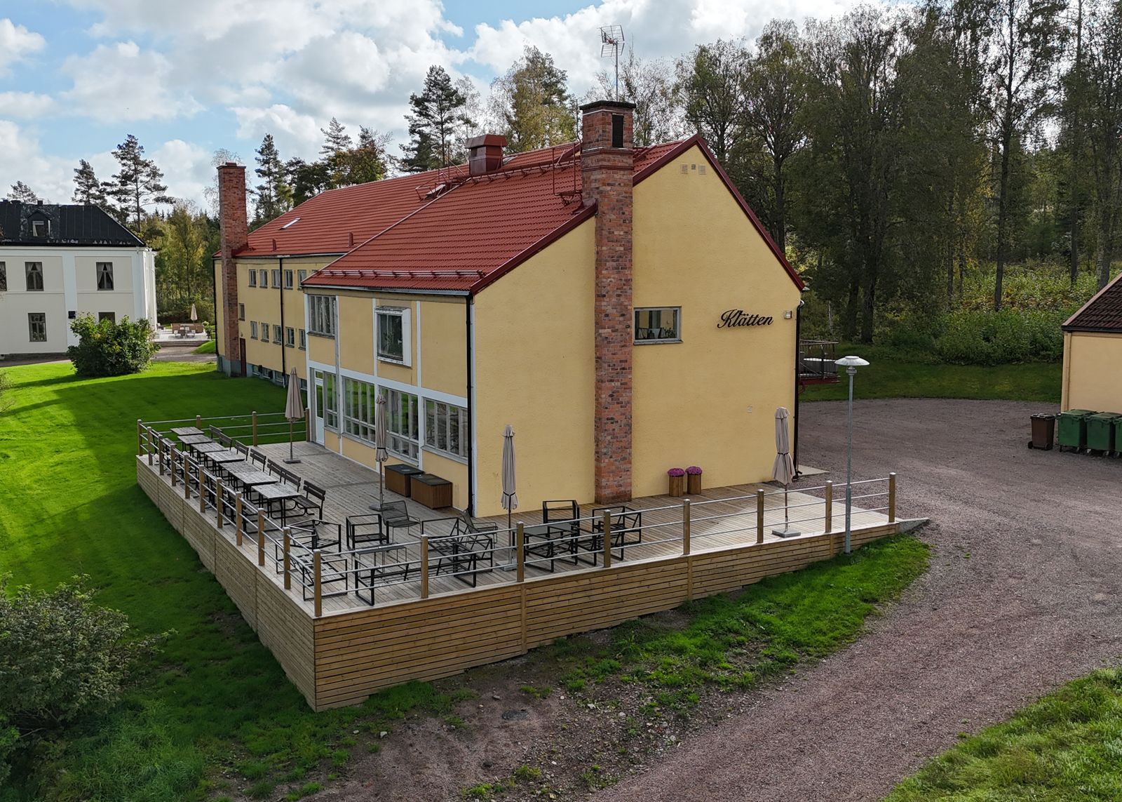 Gul byggnad med rött tak och utomhusdäck vid en grusväg i en grön lantlig miljö