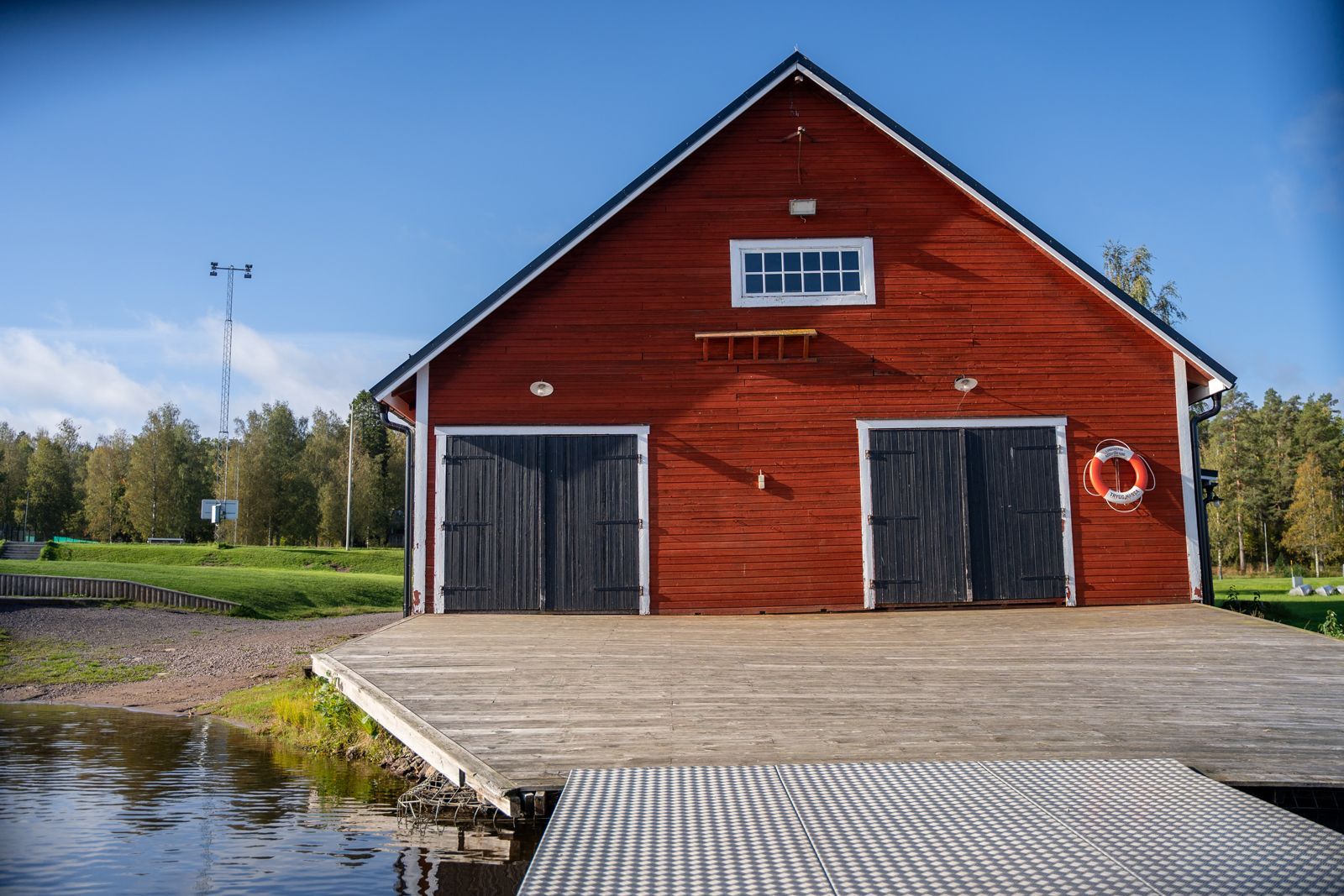 Rött träbåthus med mörka dörrar bredvid en lugn brygga vid vattnet