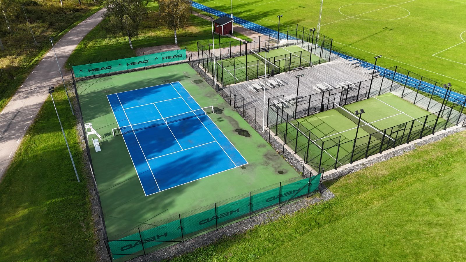 Flygfoto över blå tennisbanor bredvid inhägnade grå sportplaner på ett gräsbevuxet fält