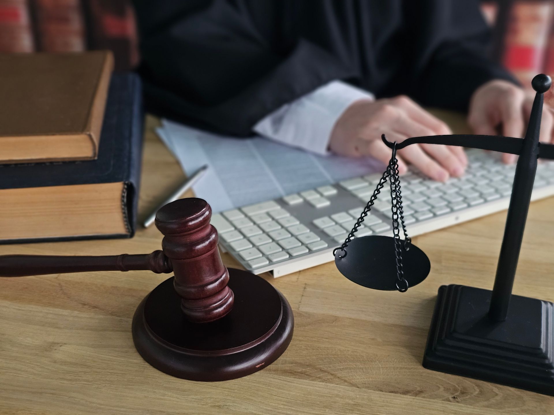 Wooden gavel and scales of justice on a desk with a person typing on a keyboard, books in background.