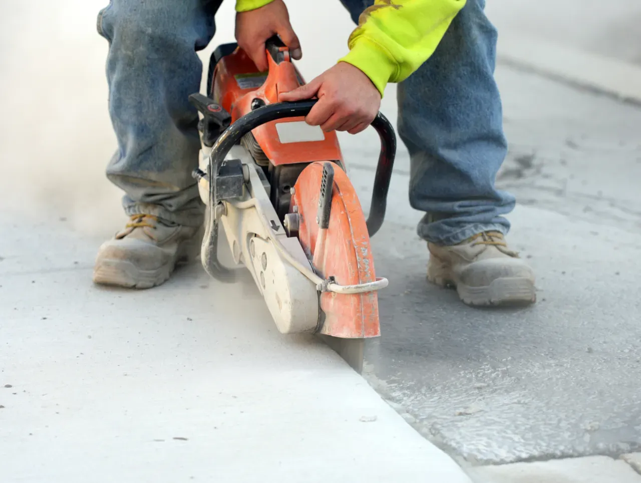 Construction worker using a saw to cut concrete. The worker wears jeans, boots, and a neon yellow shirt.