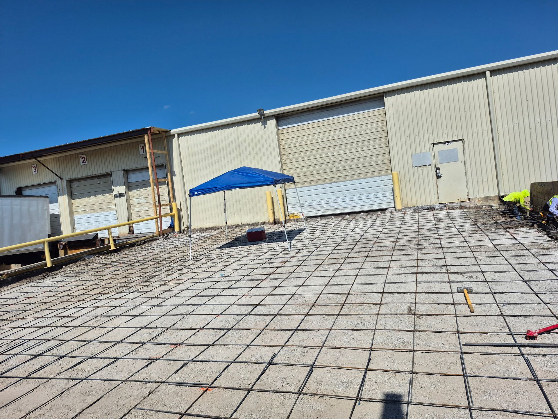 Outdoor industrial yard with a blue canopy and a large beige warehouse under a clear blue sky.