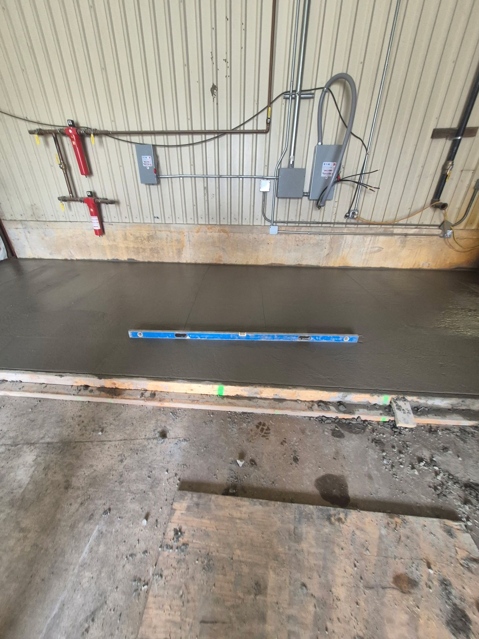Interior floor with exposed wiring and utility equipment against a corrugated wall, with a blue strip on the floor.
