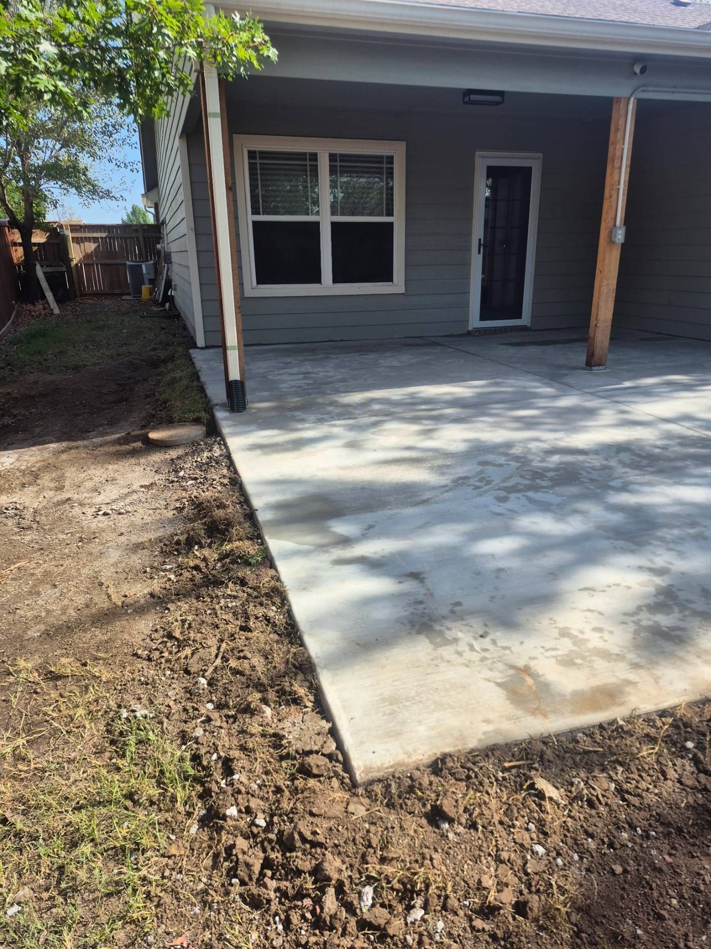 Concrete patio beside a gray house with a window, door, and wooden posts, next to a dirt yard.