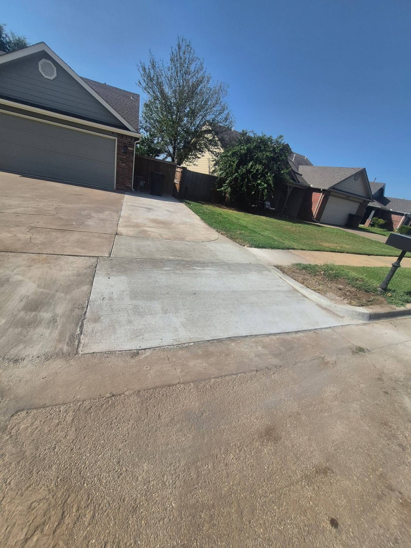 Concrete driveway on a sloped suburban street beside houses and a grassy yard under a clear blue sky