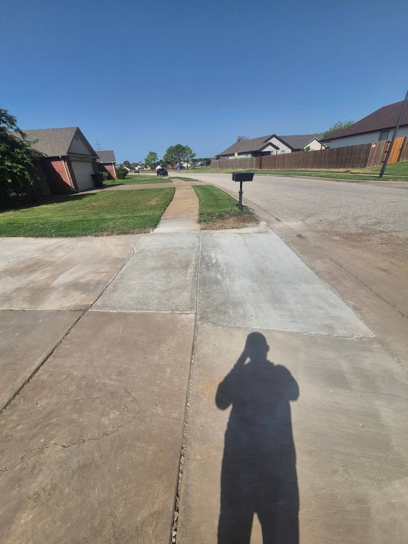 Residential street with sidewalks, driveways, mailbox, and the photographer’s shadow on the pavement under a blue sky