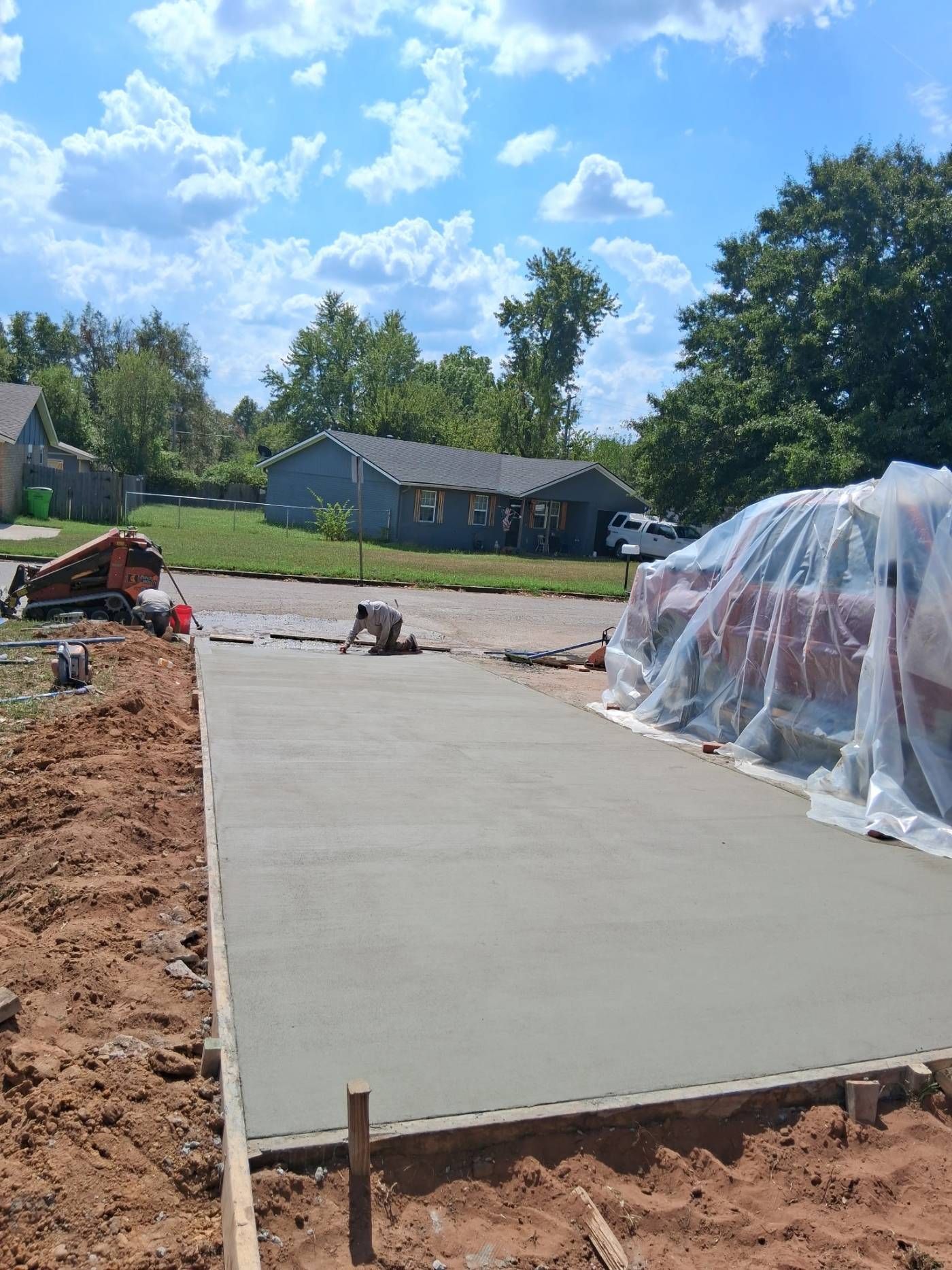 Freshly poured concrete slab at a home construction site, with plastic-covered materials nearby.