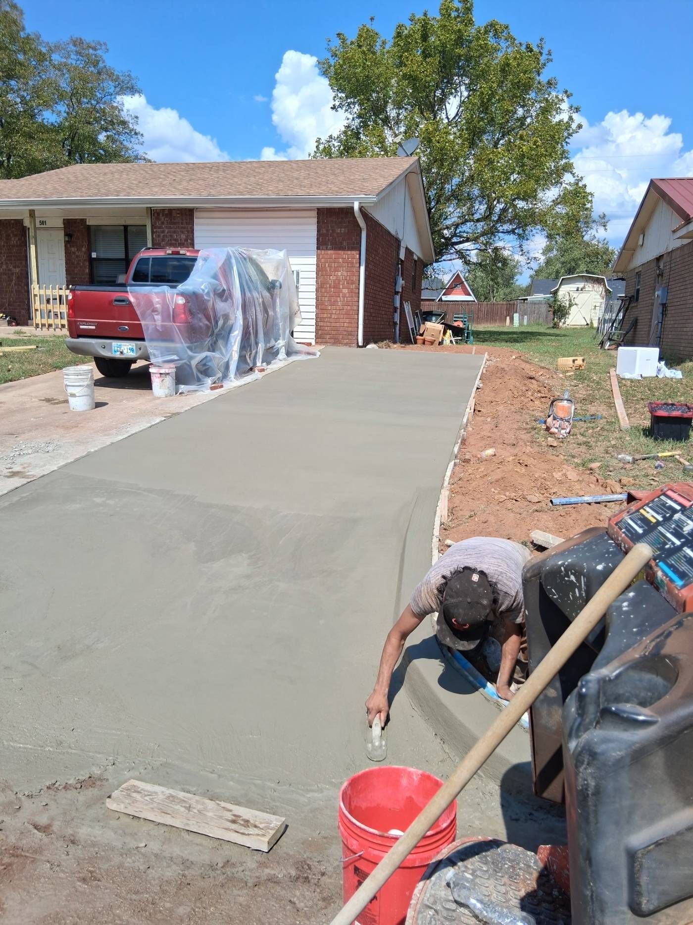 Person smoothing fresh concrete driveway beside a house, with red buckets and tools nearby.