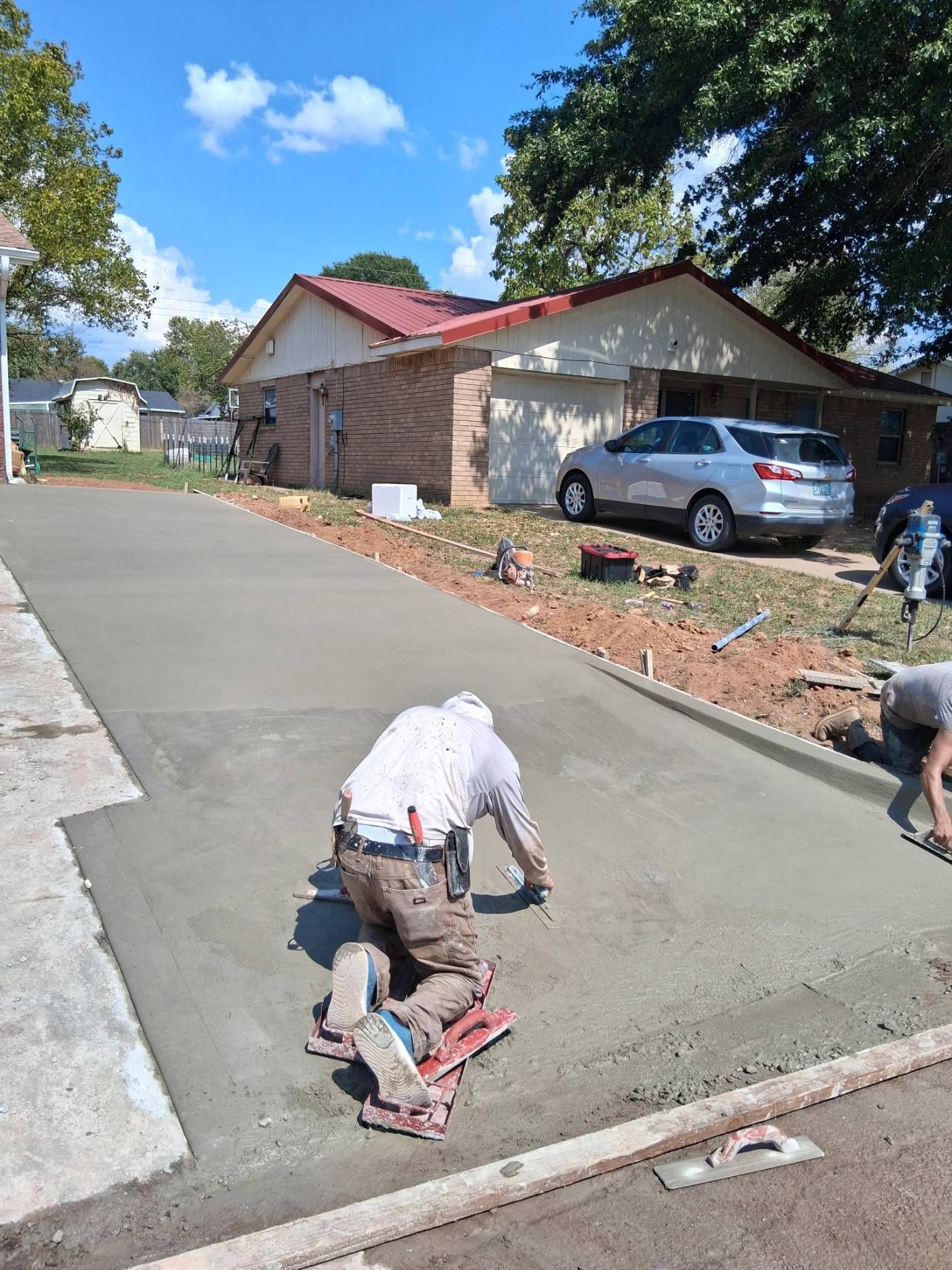 Workers smoothing fresh concrete driveway beside a house, with a parked silver car nearby.