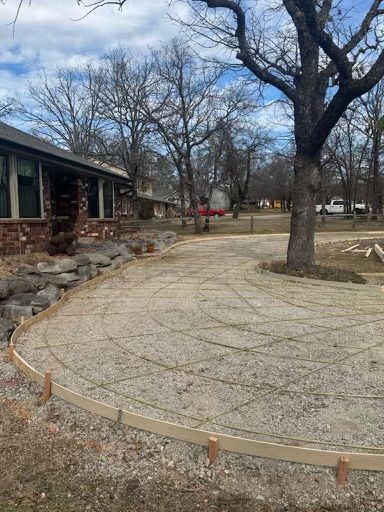 Driveway under construction with concrete forms, rebar, and gravel, next to a brick house and tree.