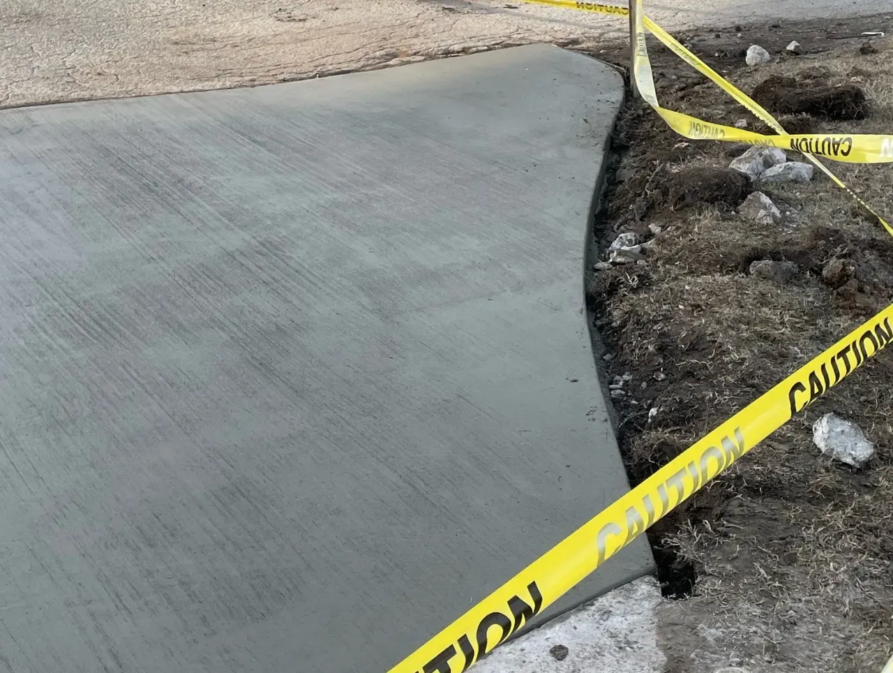 Freshly poured concrete sidewalk with caution tape along the edge, set in a dirt area.